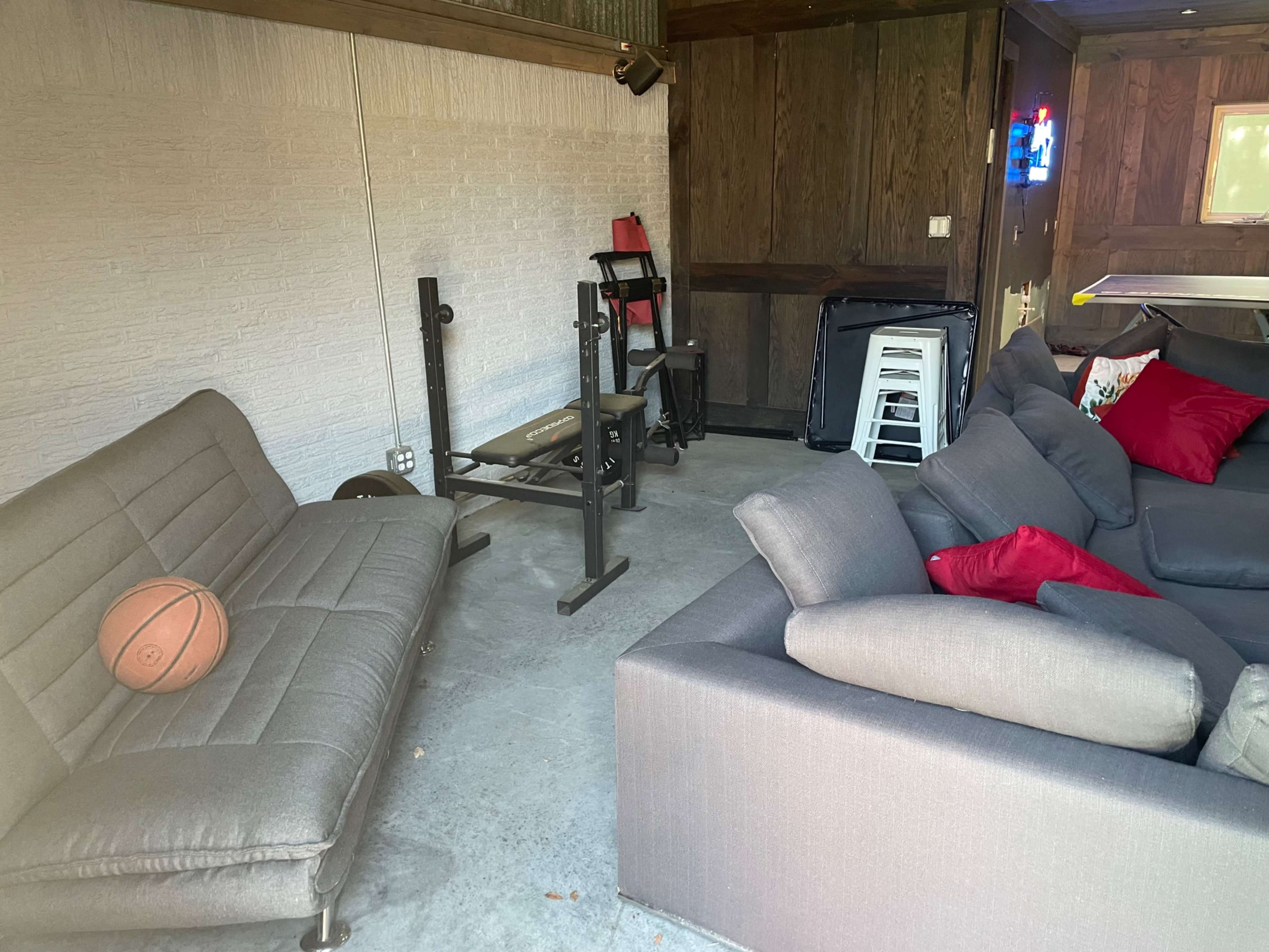 The image shows a small room with a bench press setup, a basketball on a gray couch, and a few red pillows scattered on another couch.