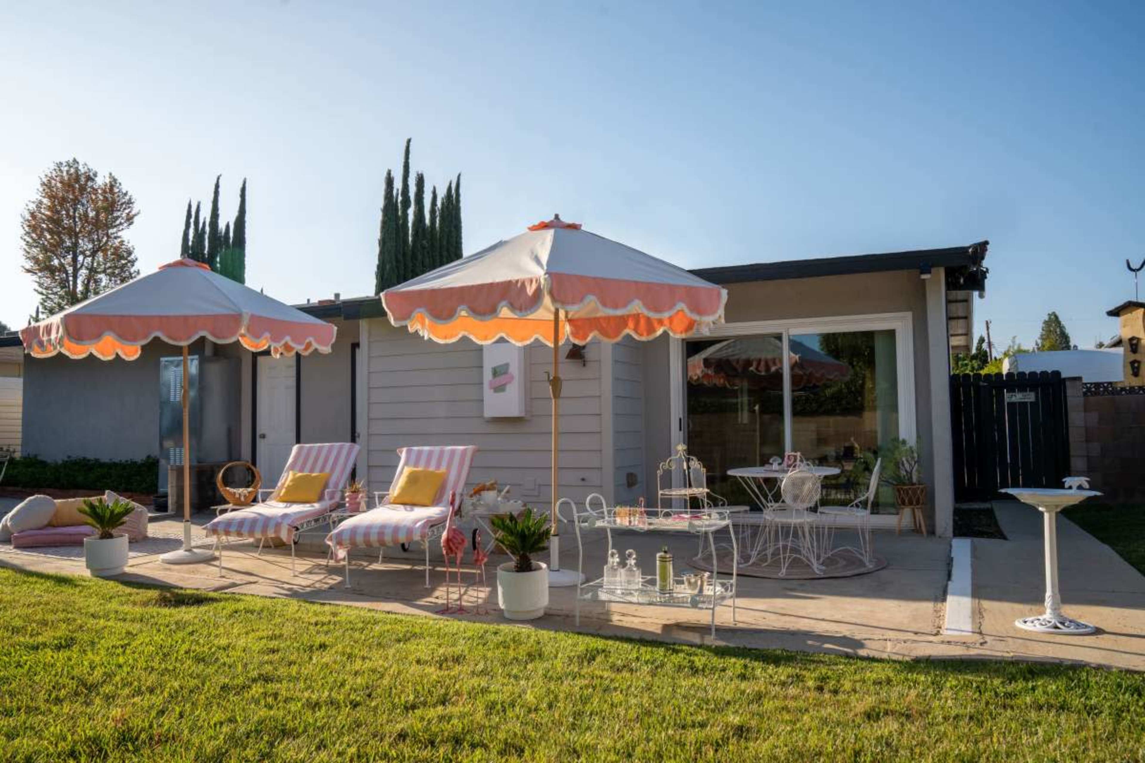 A patio area features striped lounge chairs, two large umbrellas, and a small table set against a house with a large glass door.