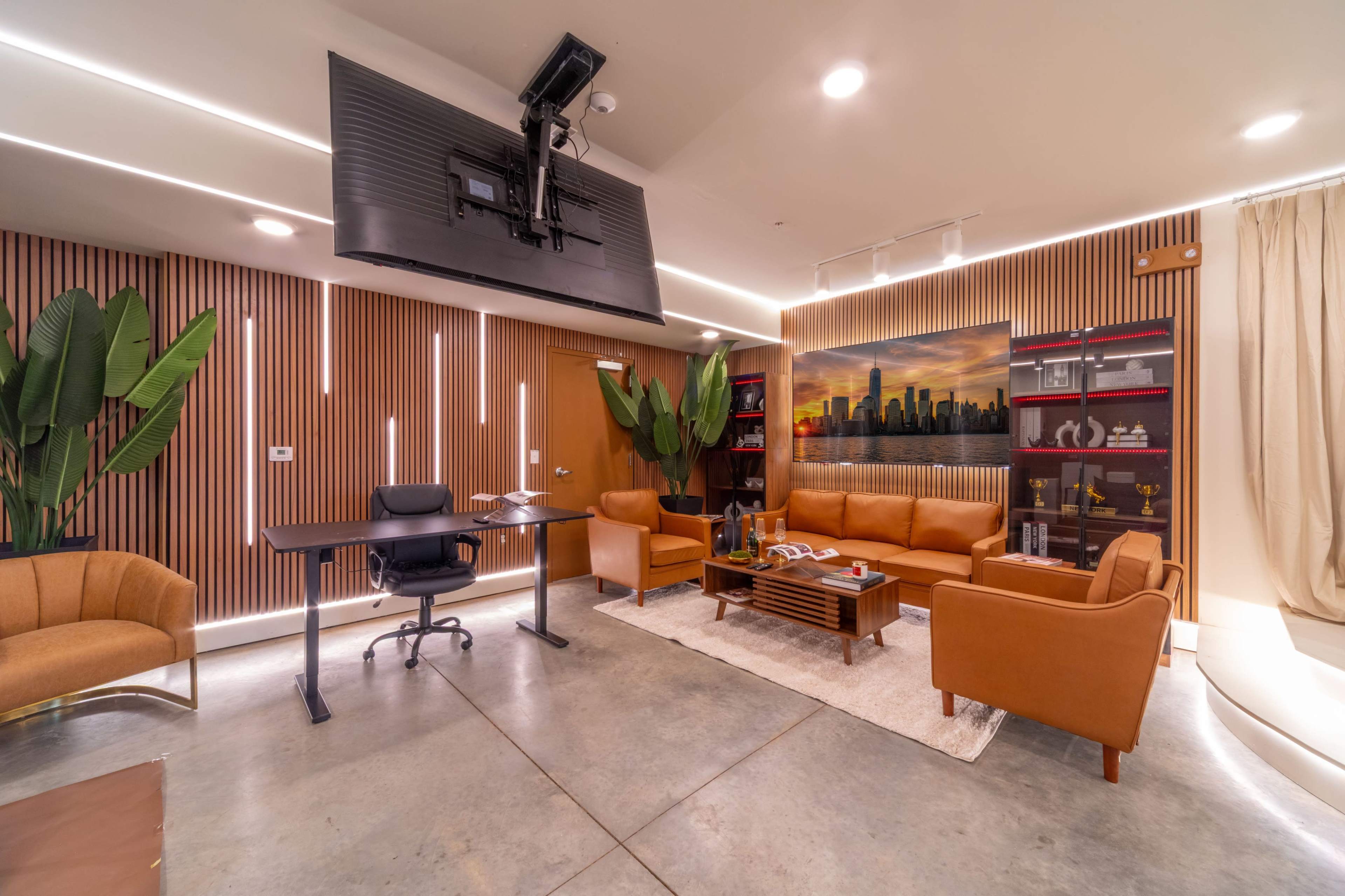 A modern office space features a brown leather sofa, a wooden desk, a large television mounted on the wall, and decorative plants, with a backdrop of a city skyline painting.