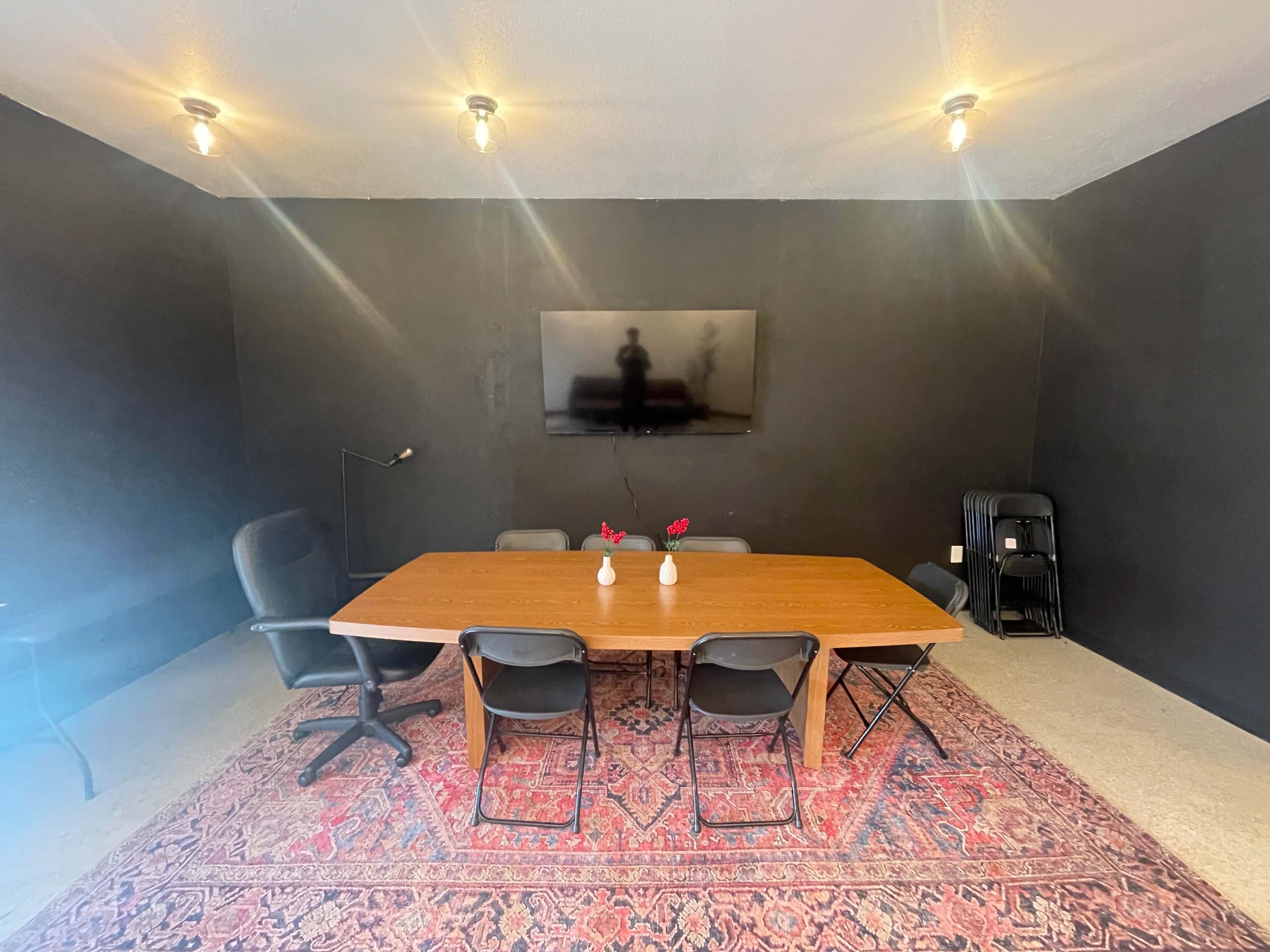 A conference room features a long wooden table surrounded by black chairs, a television mounted on the wall, and a decorative rug on the floor.