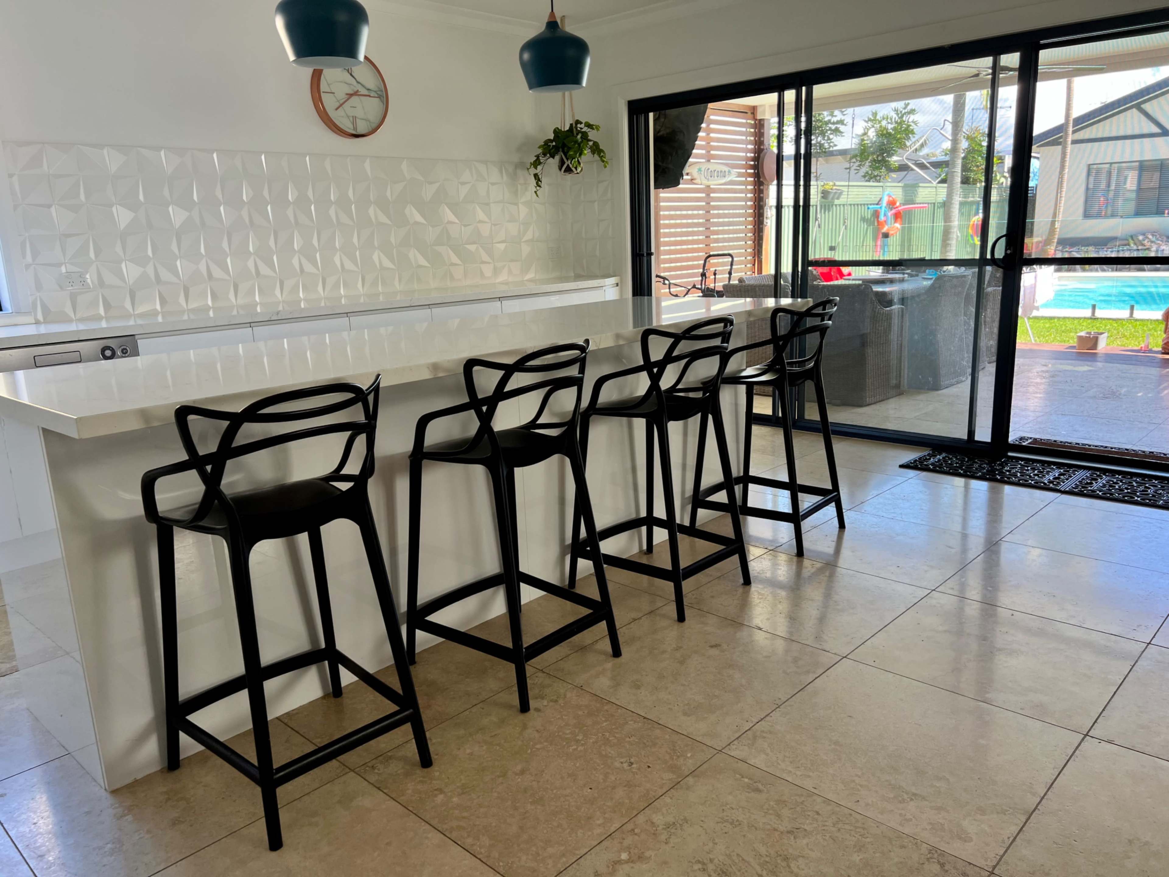 A modern kitchen features a sleek island with four black bar stools and a sliding glass door leading to an outdoor area with a swimming pool.