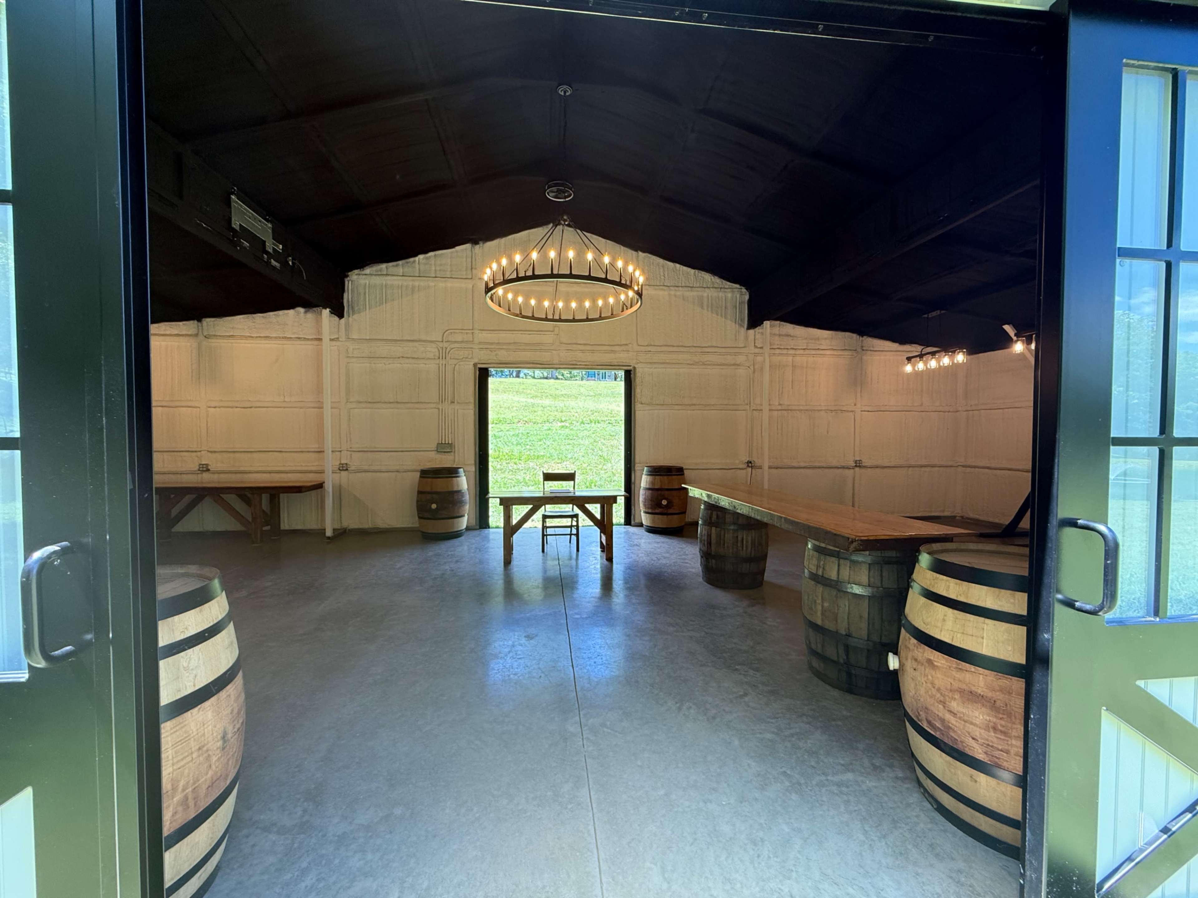 The image shows the interior of a spacious, well-lit room with wooden tables, barrels, and large doors opening to a green landscape outside.
