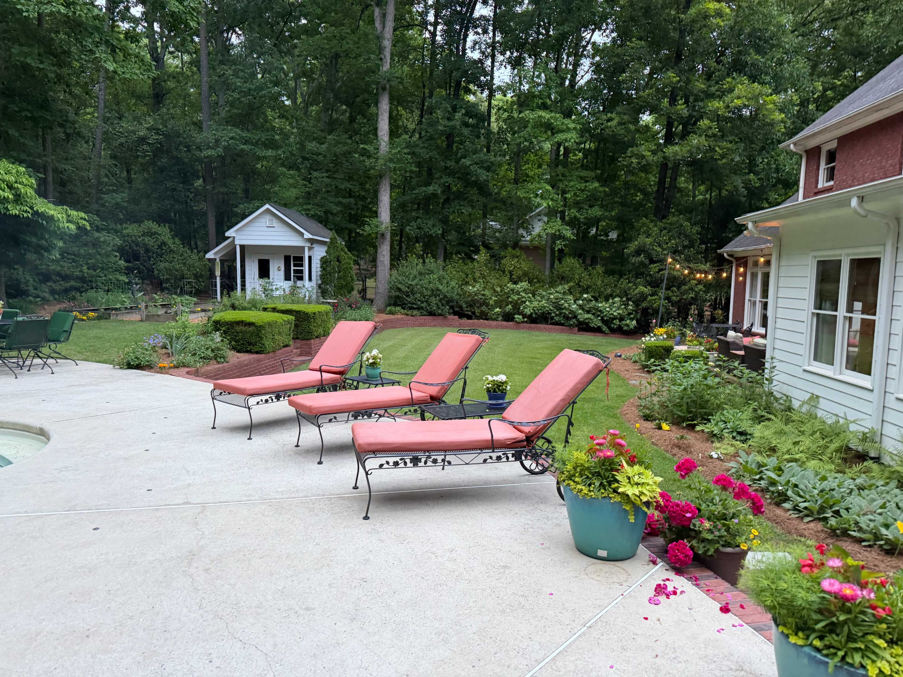 The image shows a landscaped backyard with four lounge chairs, a small shed, and various flower pots arranged around a patio area.