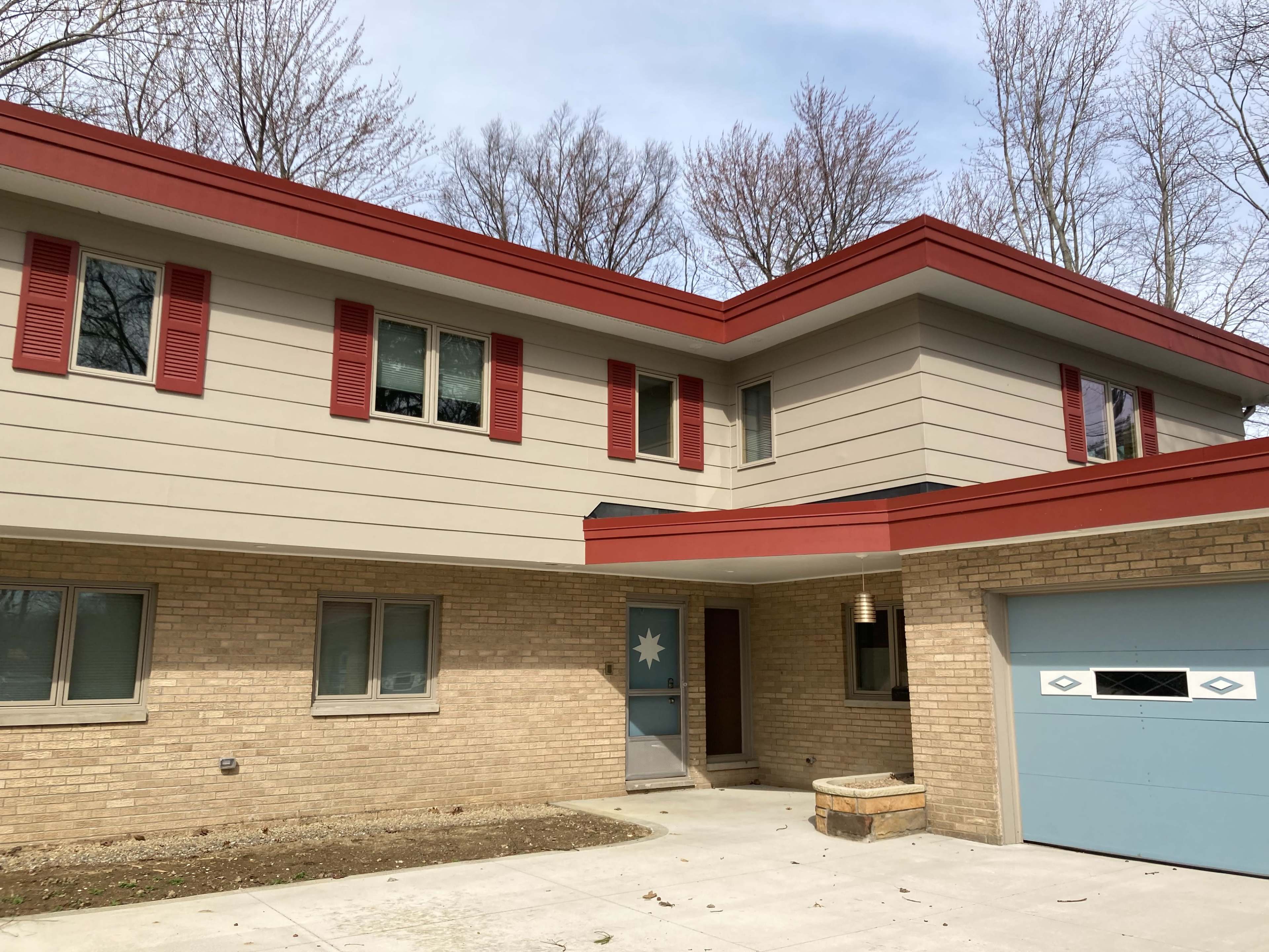 The image shows a modern two-story house with a red slanted roof, beige exterior walls, and a blue front door featuring a decorative design, set in a wooded area.
