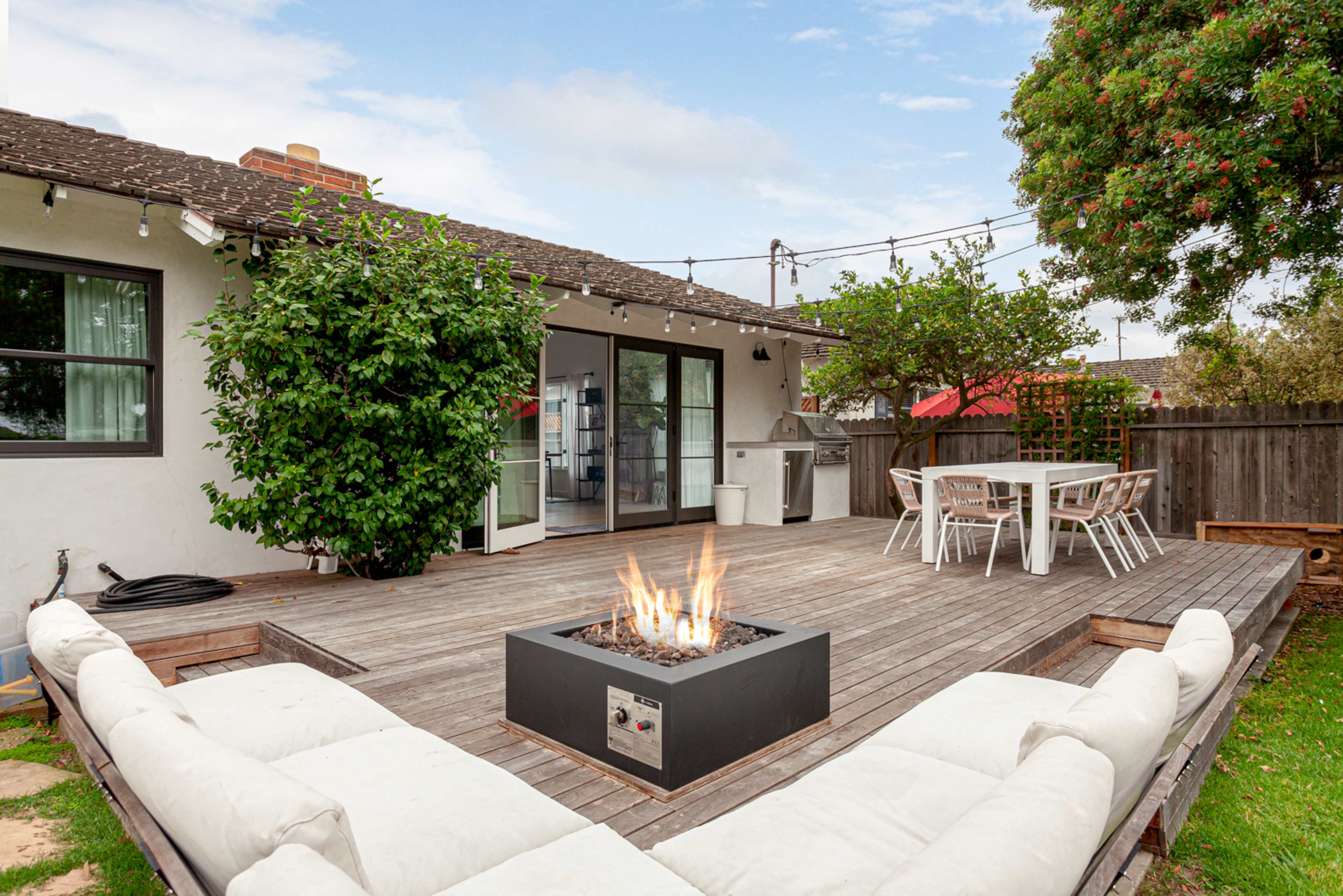 A wooden deck features a fire pit surrounded by cushioned seating, with a dining area and modern appliances in the background.
