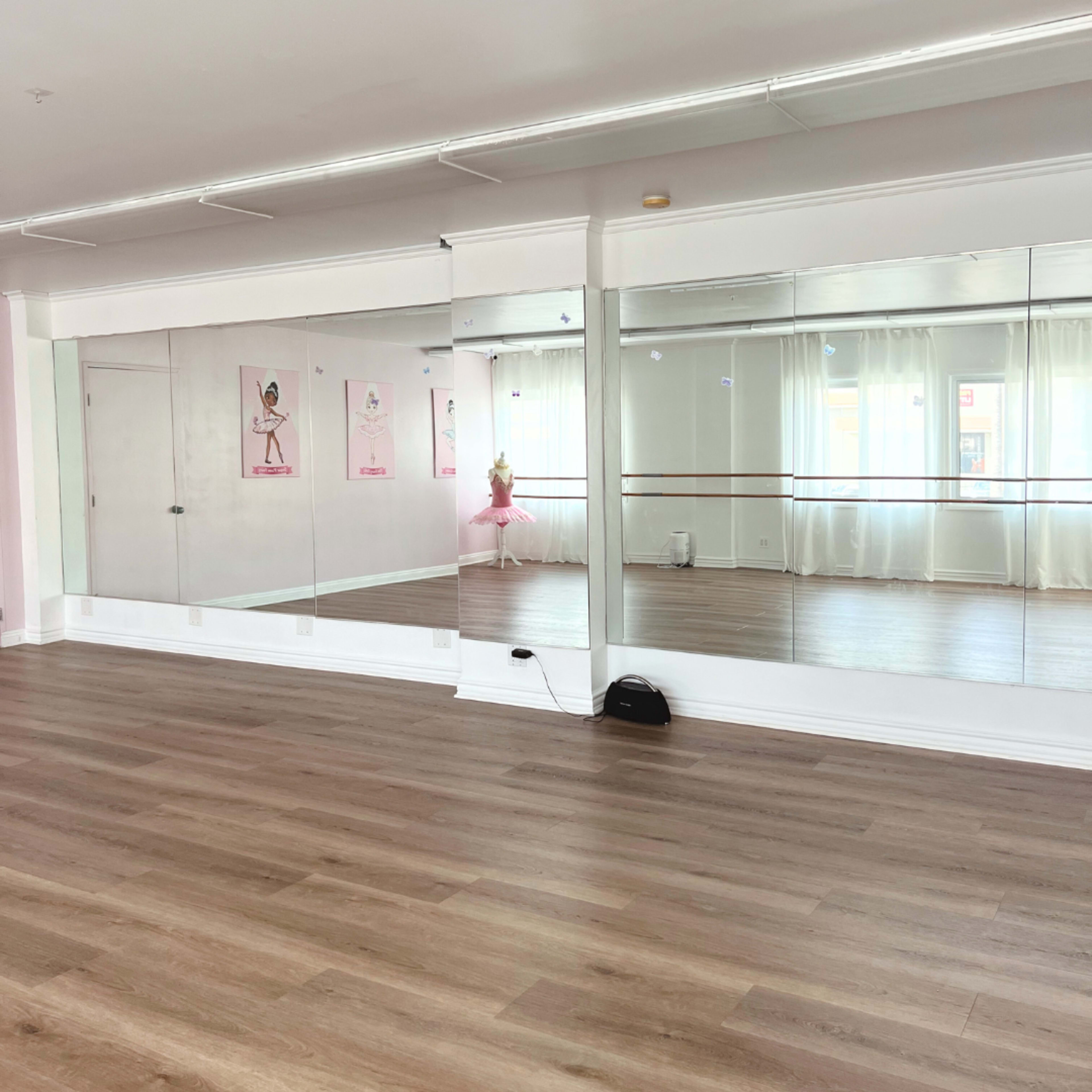 The image shows a dance studio with mirrors on one wall and a wooden floor, featuring a ballet bar and posters on the opposite wall.