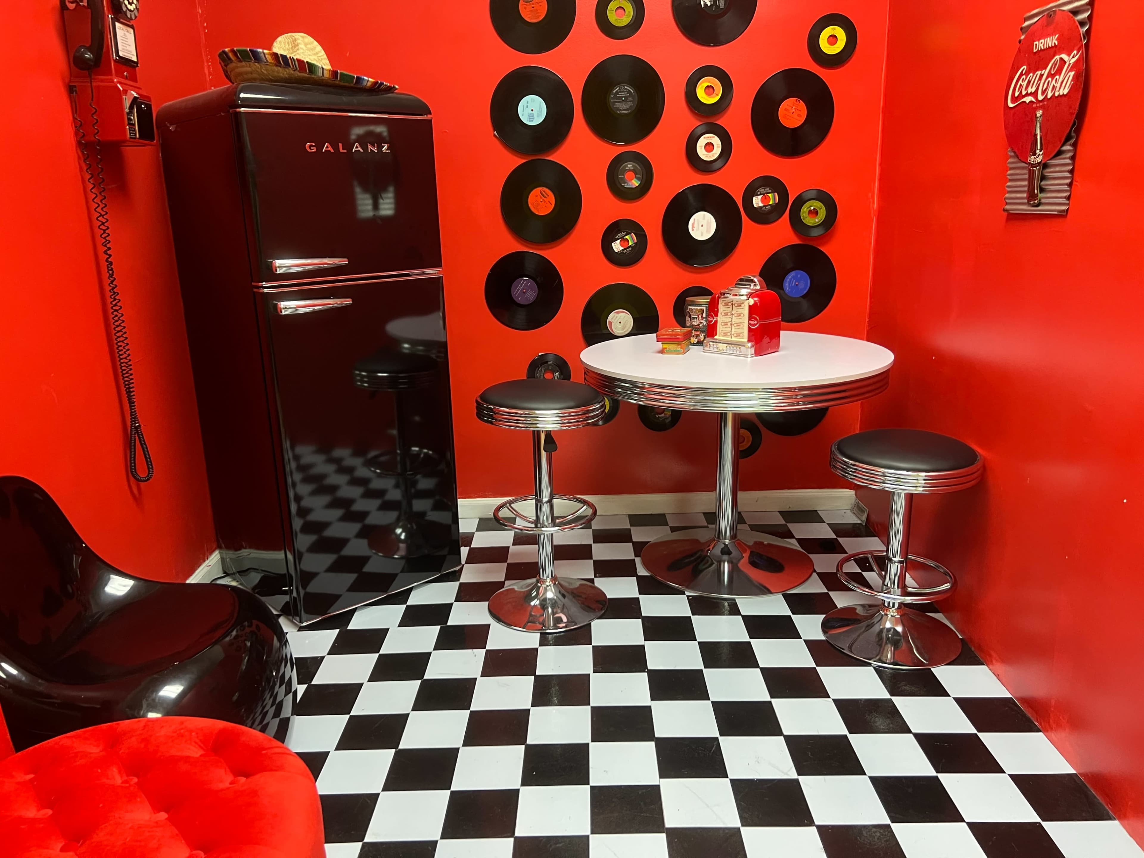 A retro-themed kitchen features a black refrigerator, a round white table with chrome stools, and a wall adorned with vinyl records against a red backdrop.