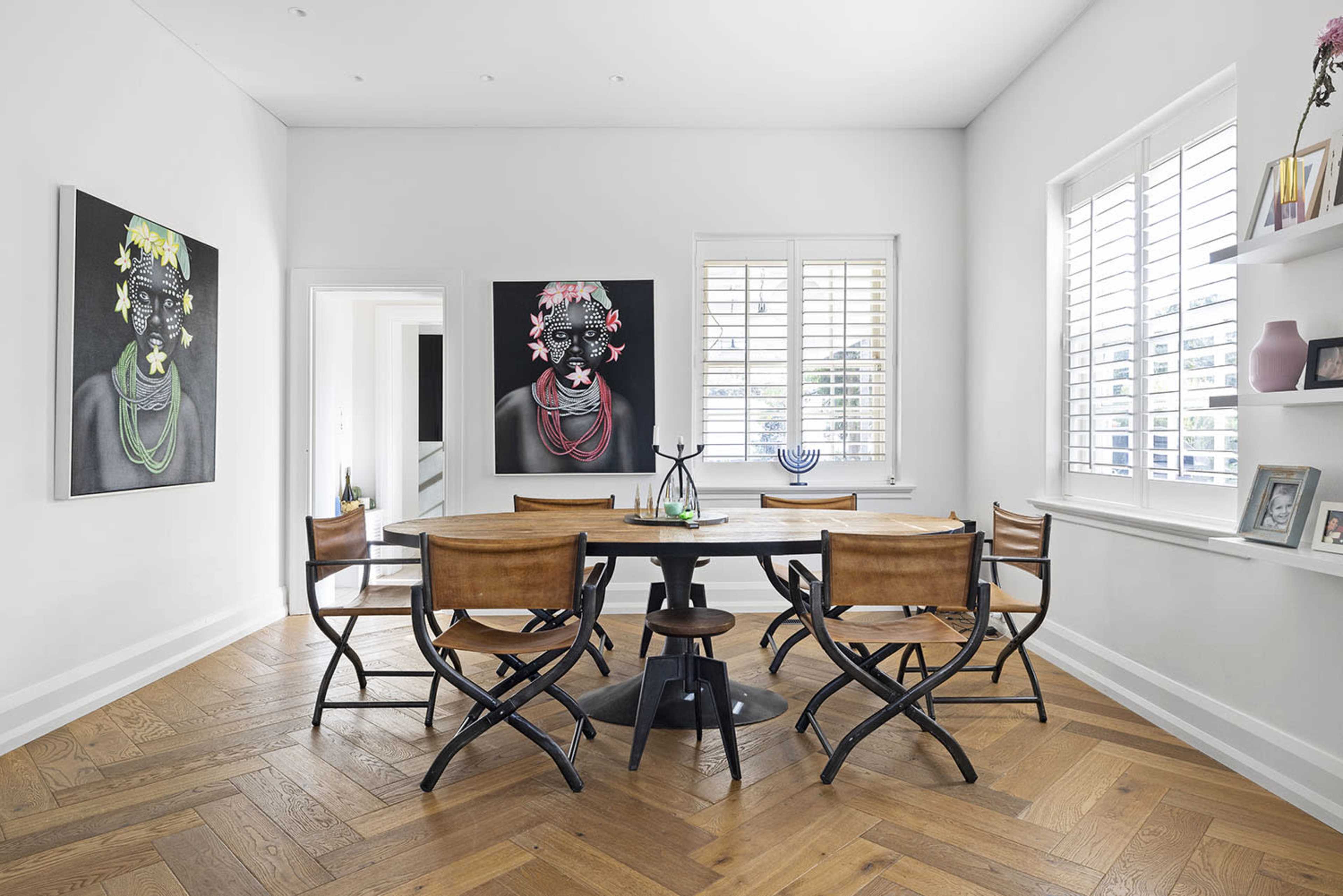 A modern dining area featuring a round table surrounded by several chairs, with two large framed artworks on the wall and a shelf displaying various decor items.