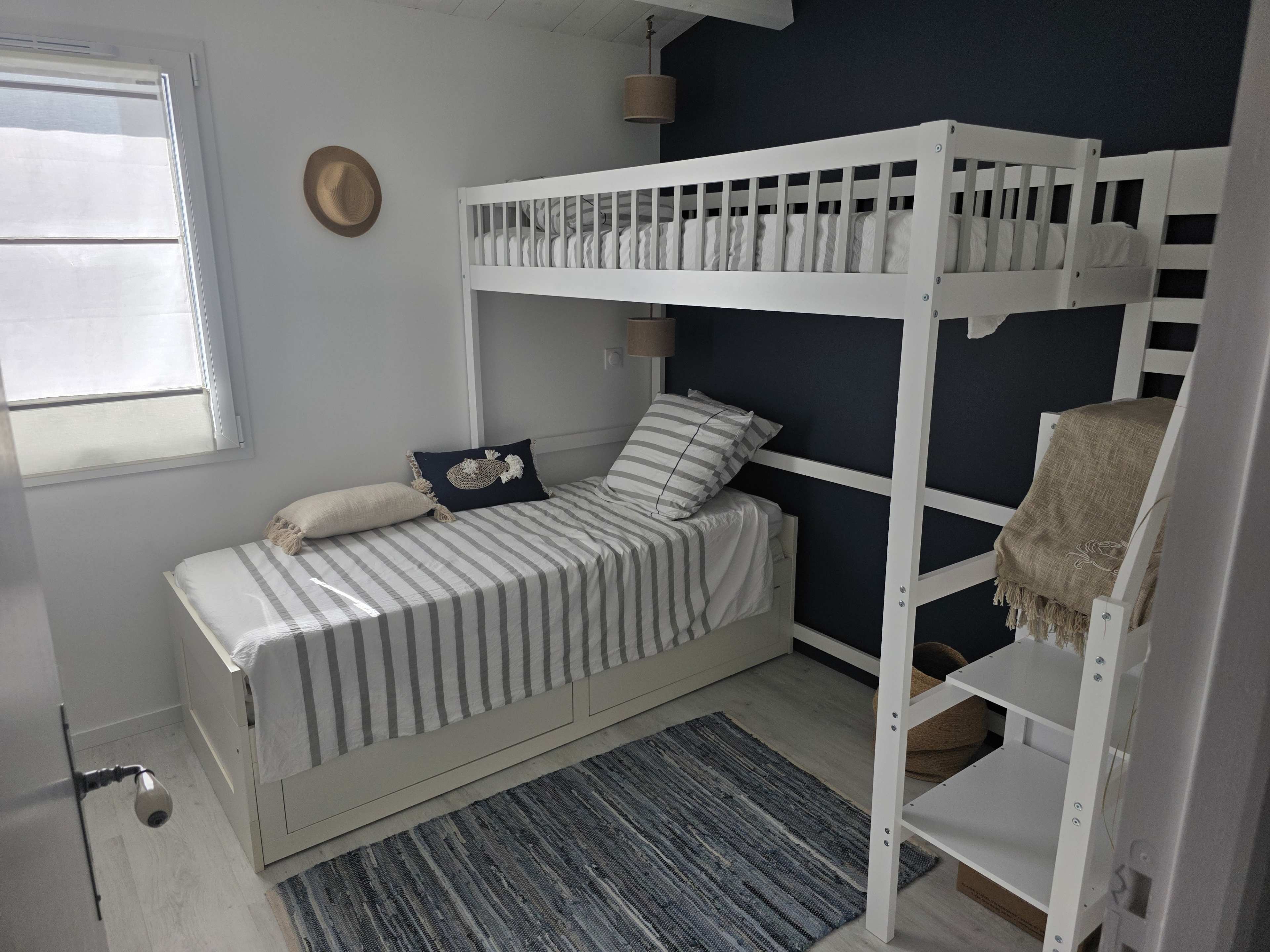 The image shows a small bedroom featuring a bunk bed with a single bed on the bottom and a lofted bed on top, alongside a small bedside table and a woven area rug.