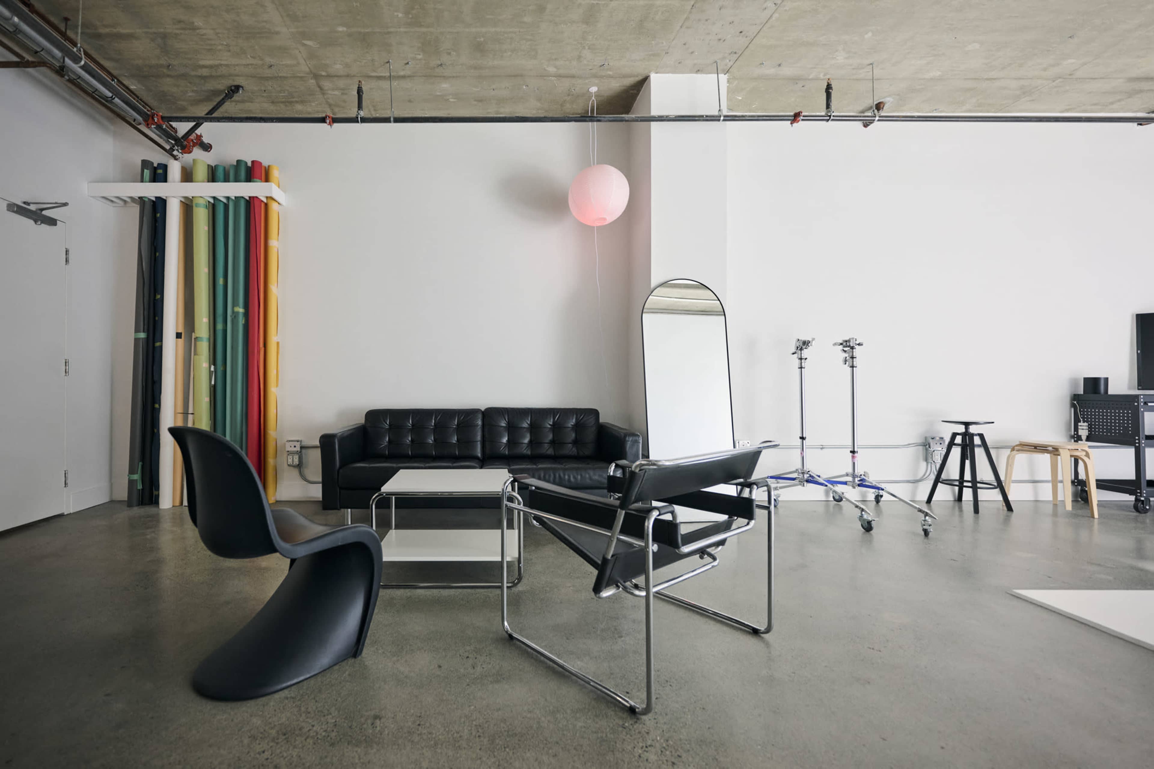 The image shows a modern, minimalist studio featuring a black leather sofa, a mirrored standing mirror, a glass coffee table, and a sleek black chair against a backdrop of colorful rolls of material.