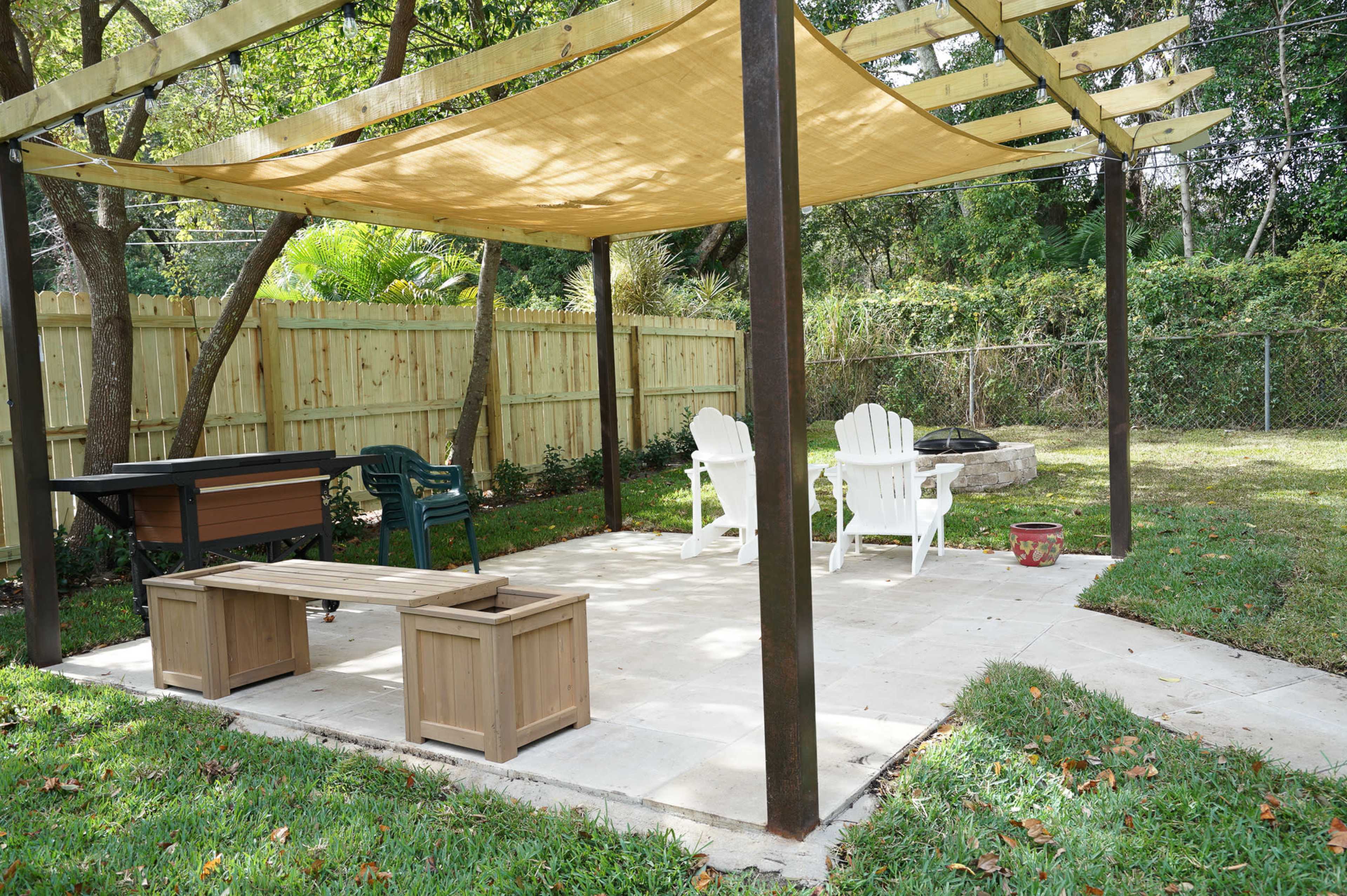 The image shows a shaded outdoor patio area with two white Adirondack chairs, a wooden bench, and a stone fire pit surrounded by grass.