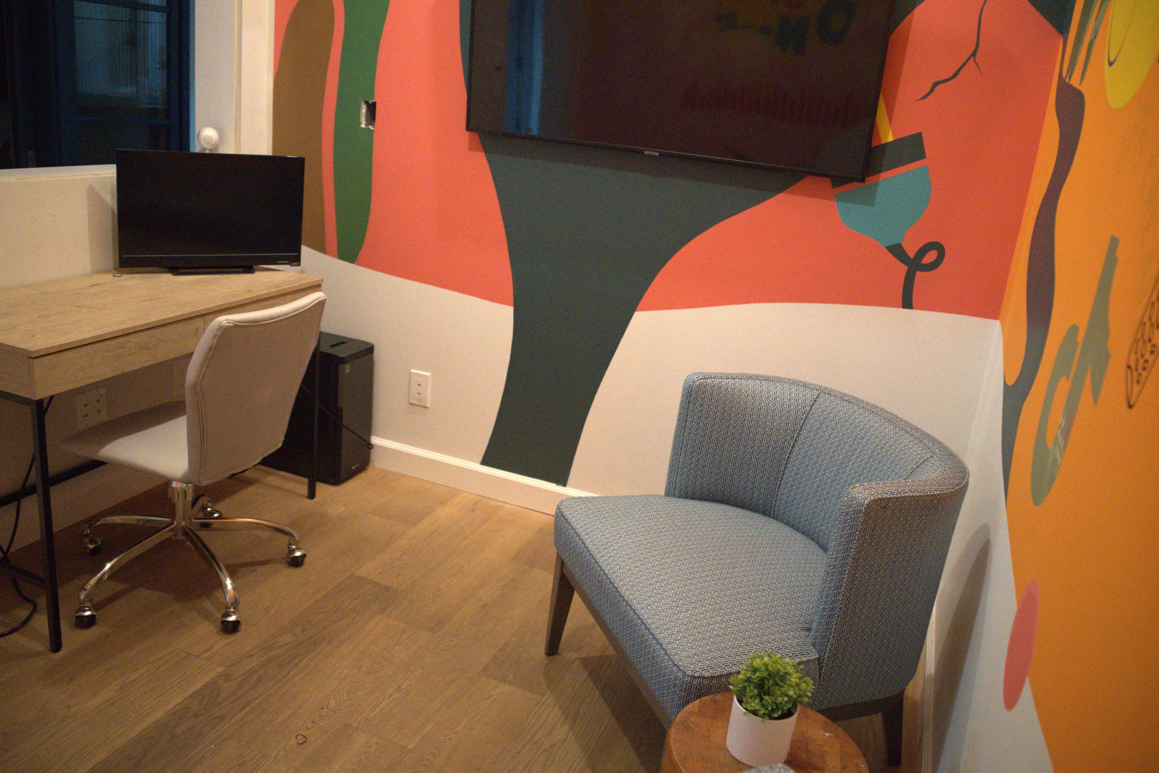The image shows a small office space featuring a wooden desk with a computer monitor, a light blue accent chair, and a colorful mural on the wall.