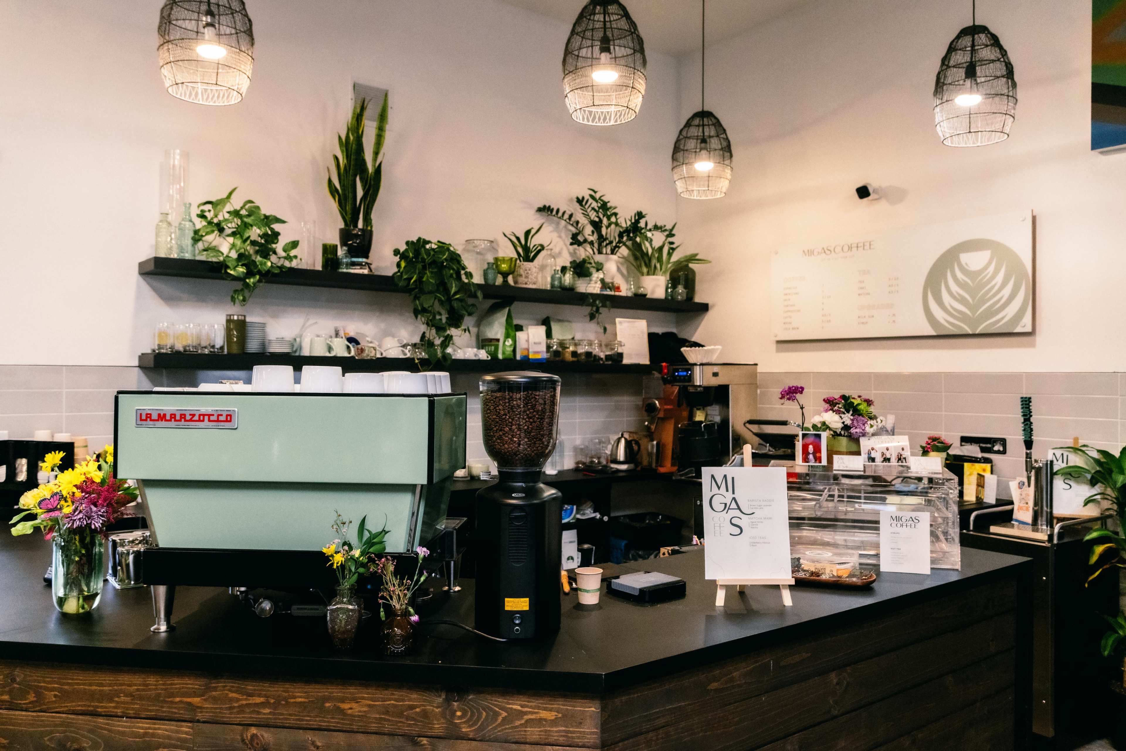 The image shows a modern coffee shop interior with a green espresso machine, a wooden counter, various plants, and a menu on the wall.