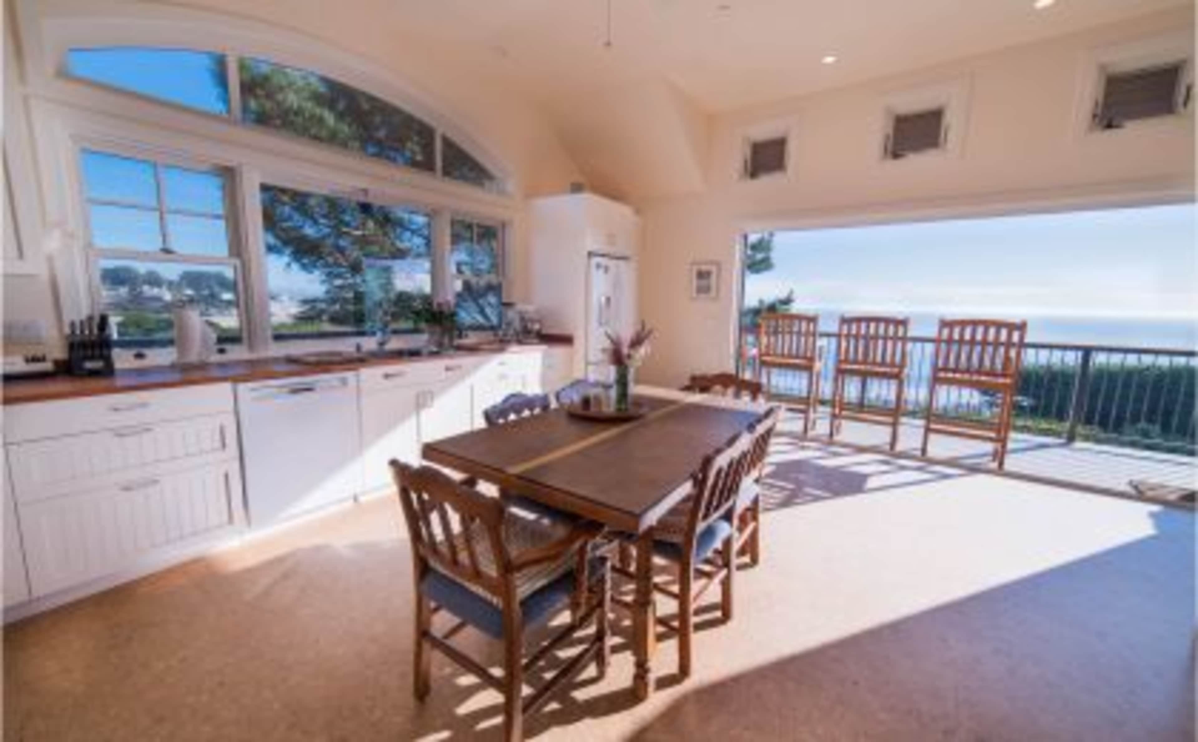 The image shows a bright kitchen and dining area featuring a large wooden table surrounded by chairs, with a view of the outdoors visible through large windows.