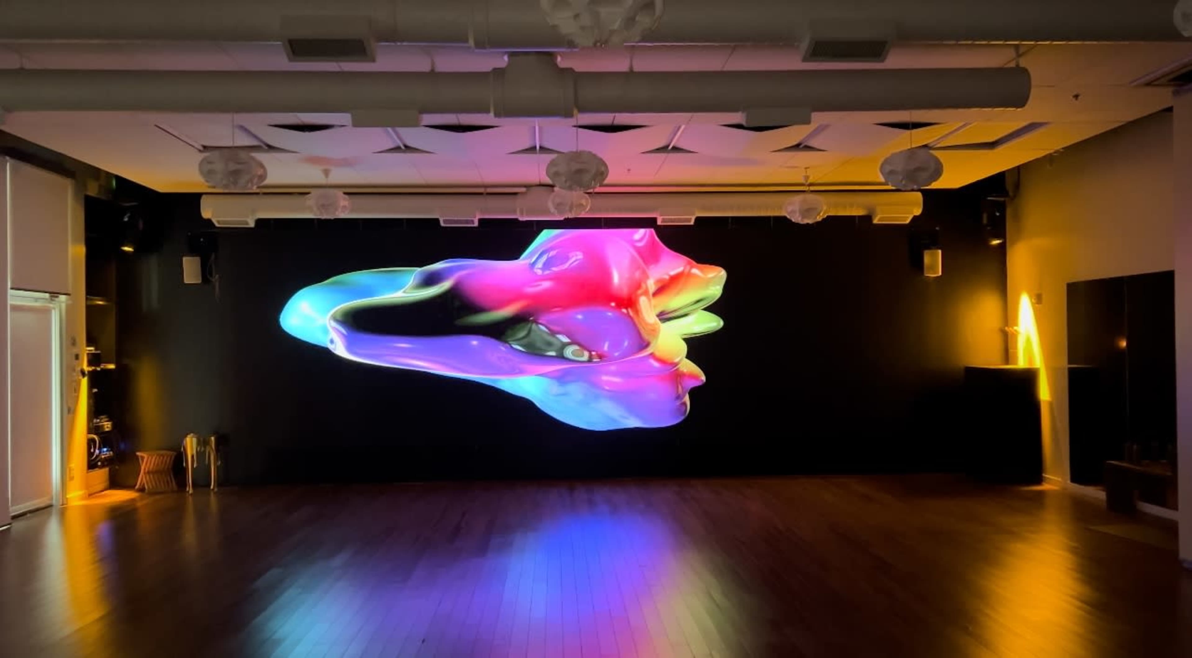 A large, colorful 3D projection of an abstract shape is displayed on a black wall in a dimly lit room with wooden flooring.