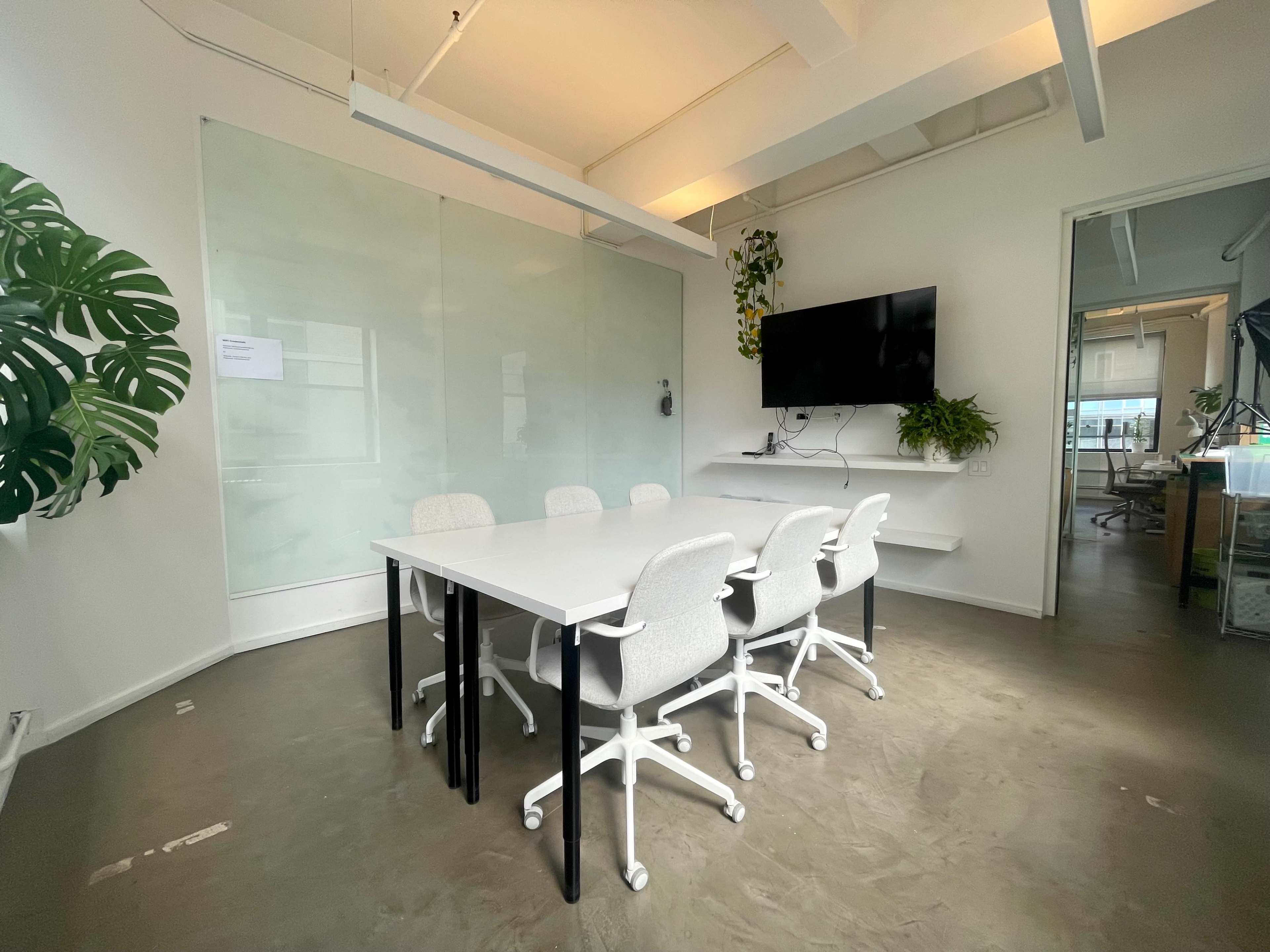 A minimalist conference room features a white table surrounded by six grey chairs, with glass walls and a wall-mounted screen.
