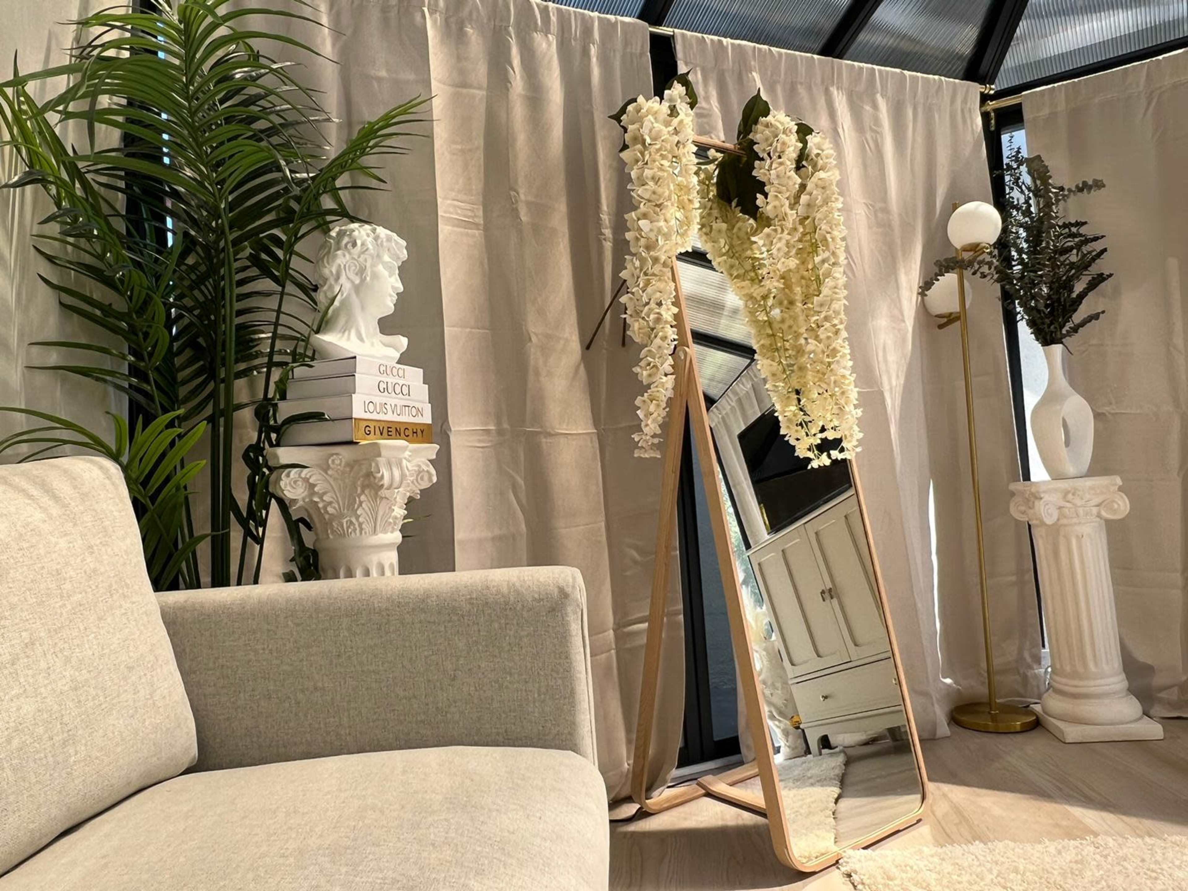 A large mirror stands against a wall adorned with white curtains, flanked by decorative plants and a vintage-style statue.