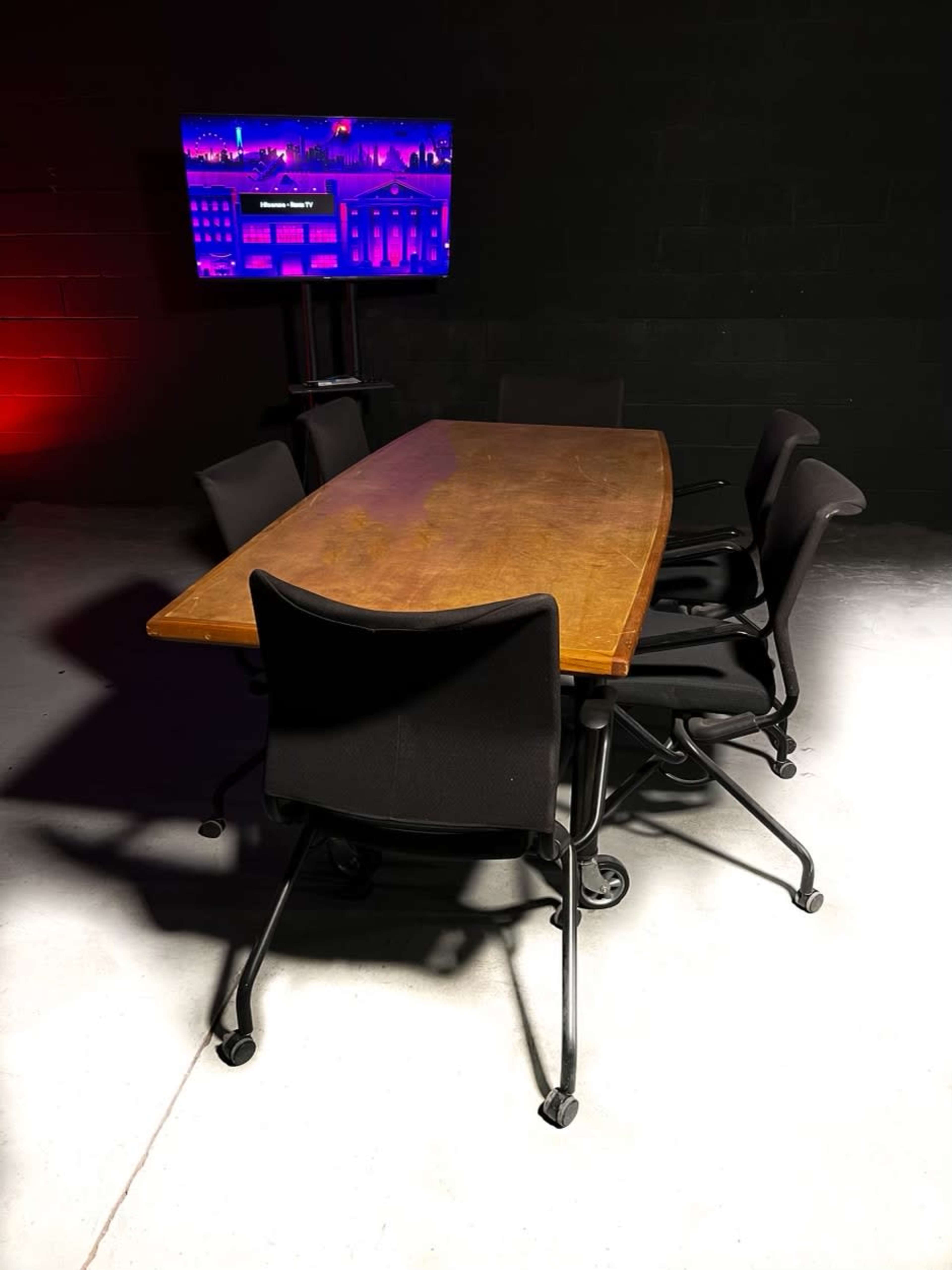 A long wooden conference table is surrounded by six black rolling chairs, with a television screen mounted on the wall behind it.