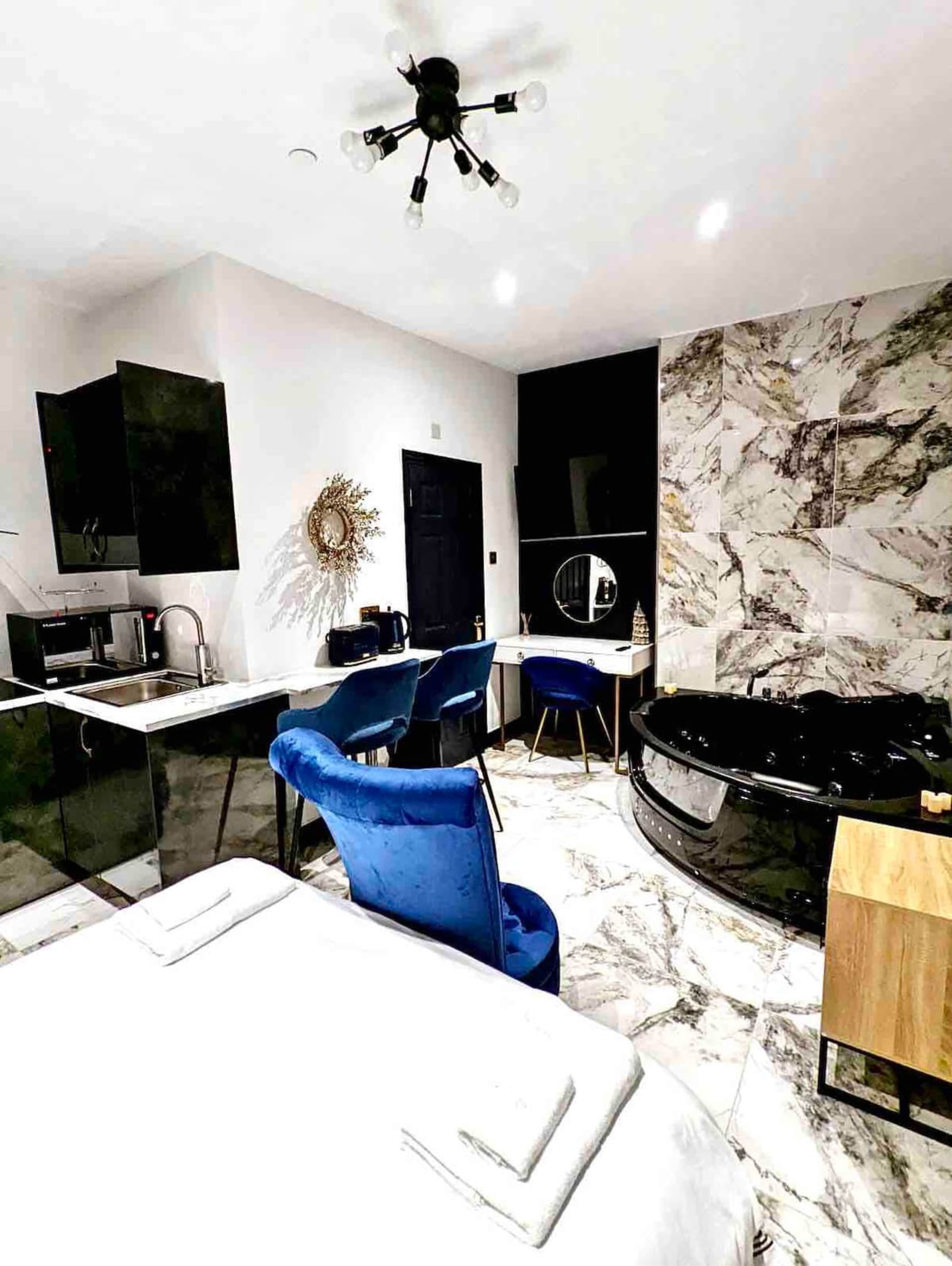 The image shows a modern hotel room featuring a bed, a kitchen area with black cabinetry, a small dining table with blue chairs, and a black whirlpool tub.