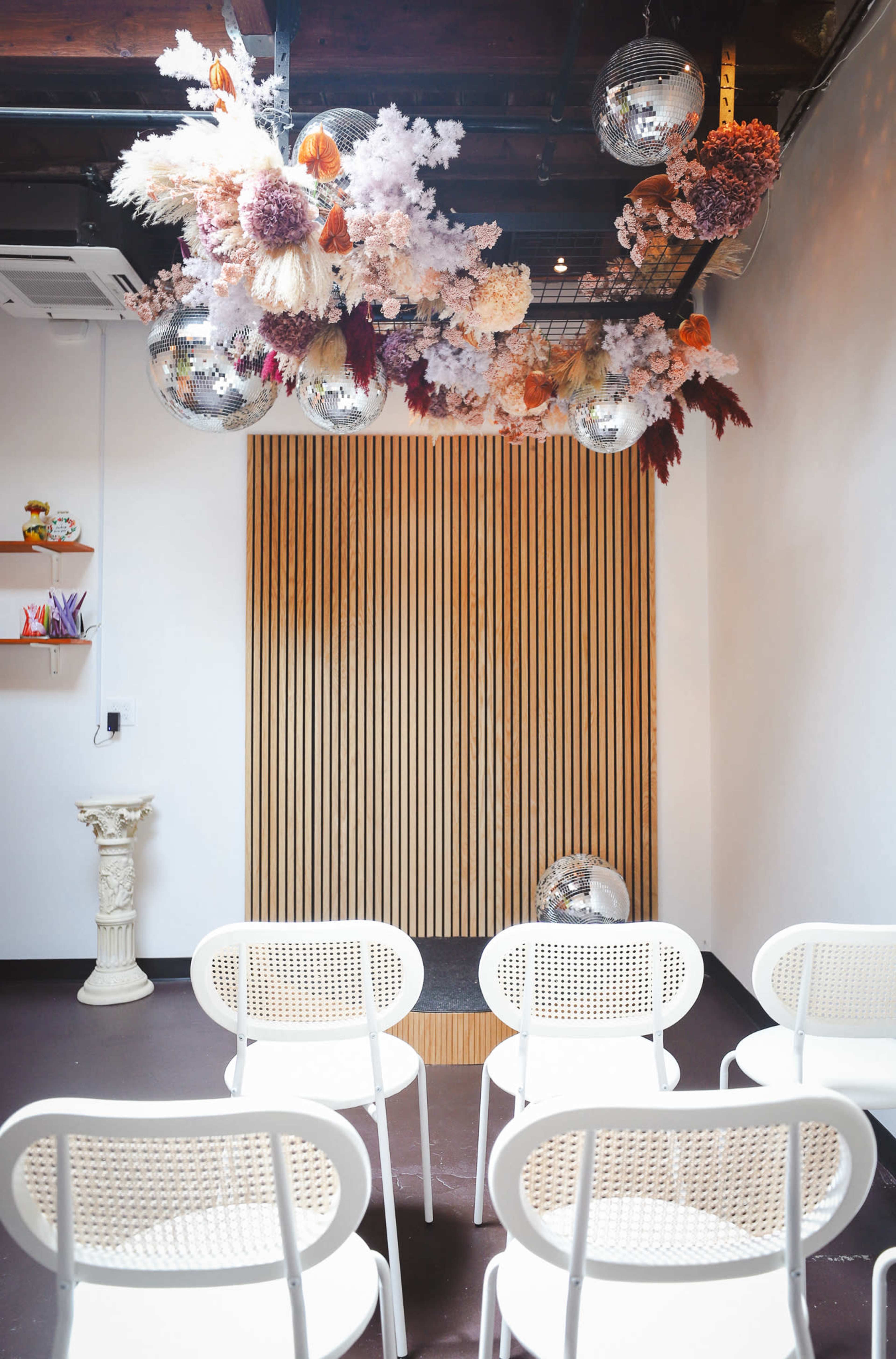 A room with six white chairs arranged in front of a wooden wall, decorated with colorful floral installations and hanging disco balls.