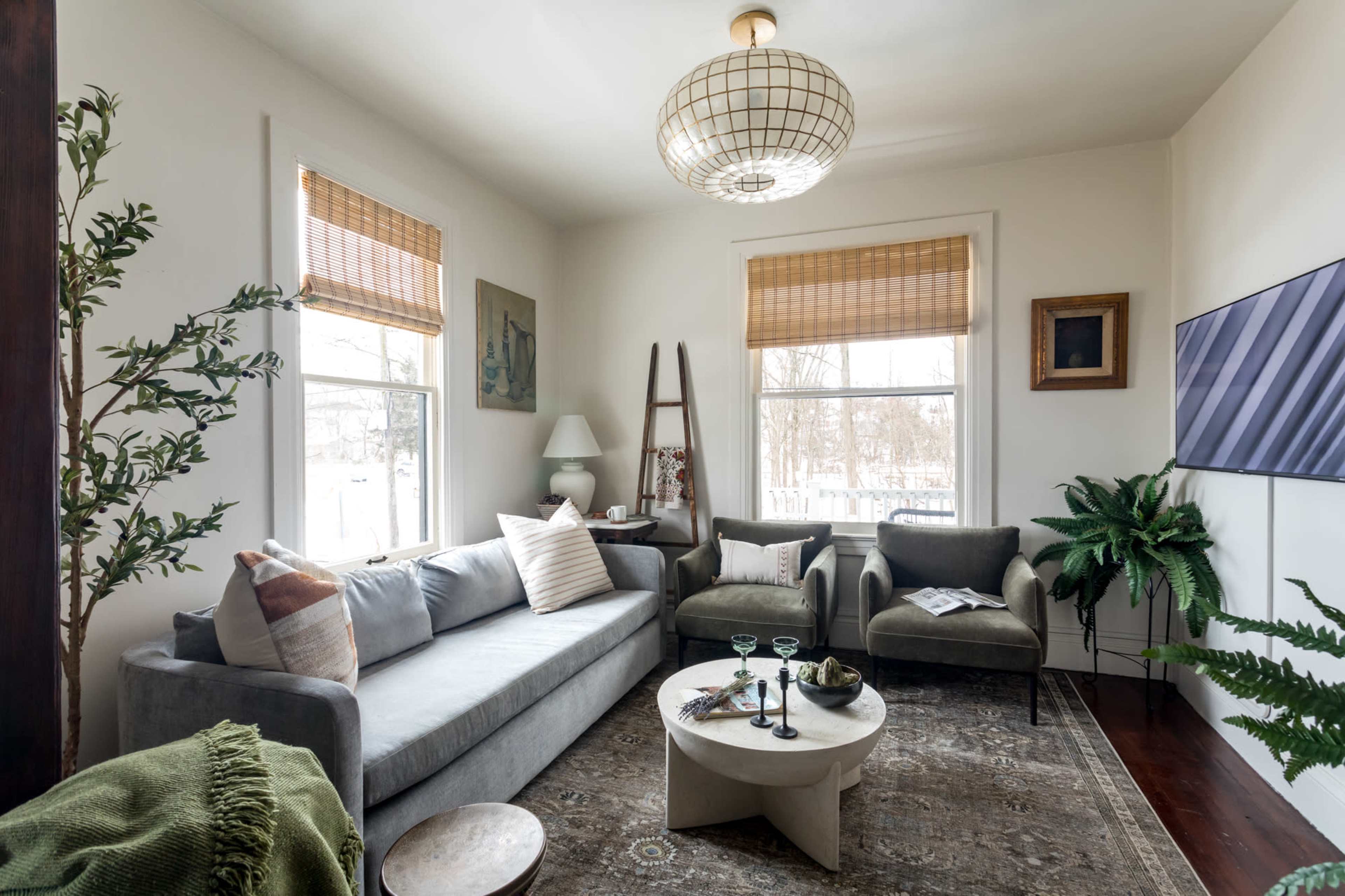 A cozy living room features a gray sofa, two green chairs, a round coffee table, and large windows with bamboo shades, surrounded by plants.