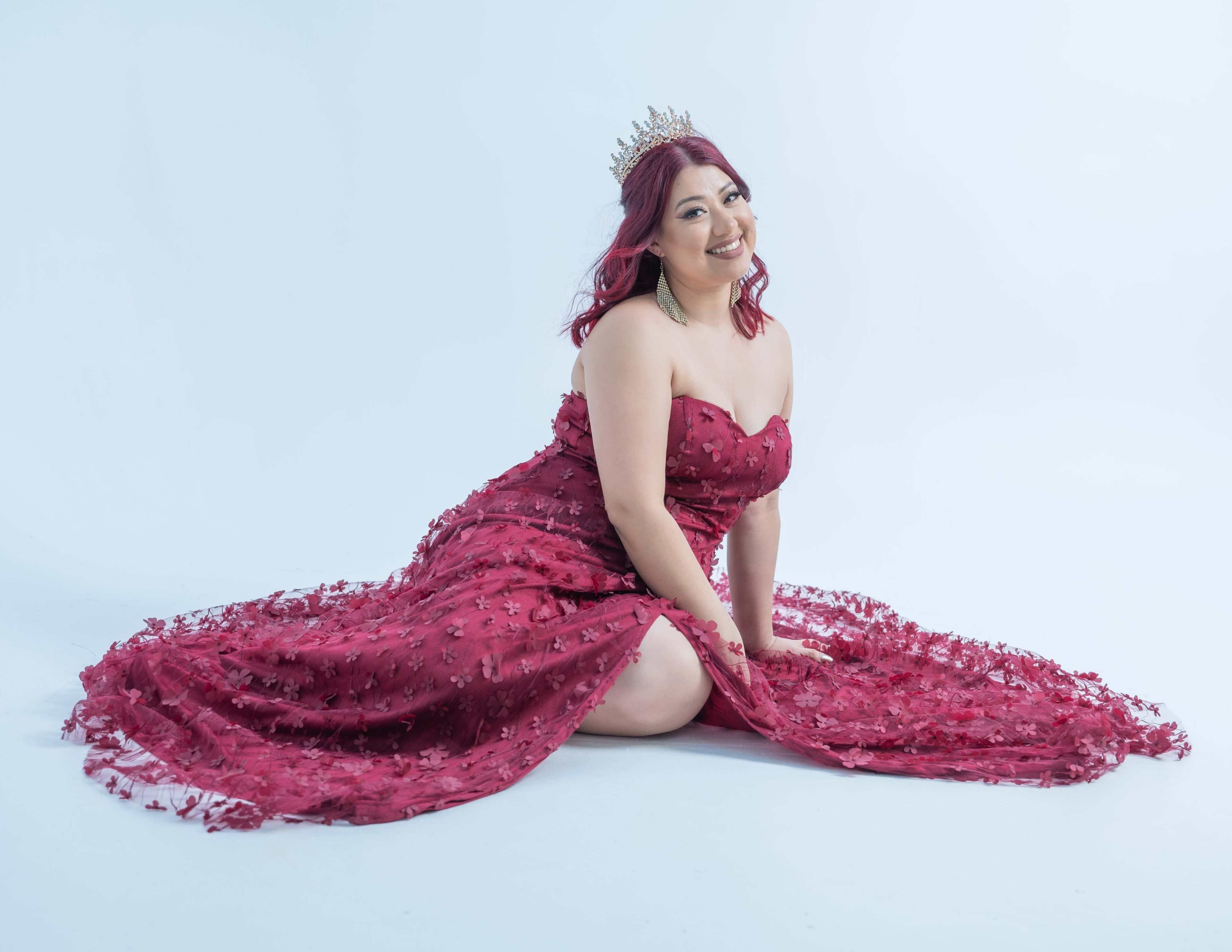 A woman in a red, elaborately designed gown kneels on a white backdrop while wearing a crown.