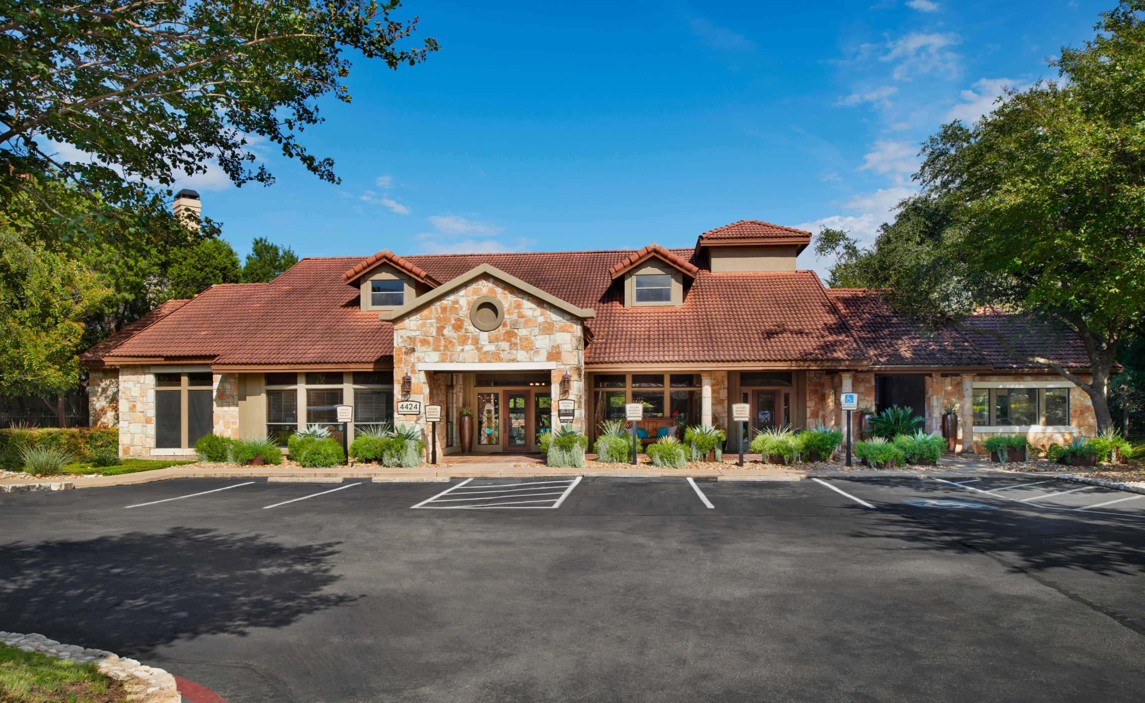 A large stone-clad building with a red tile roof, surrounded by landscaped greenery and a paved parking area.