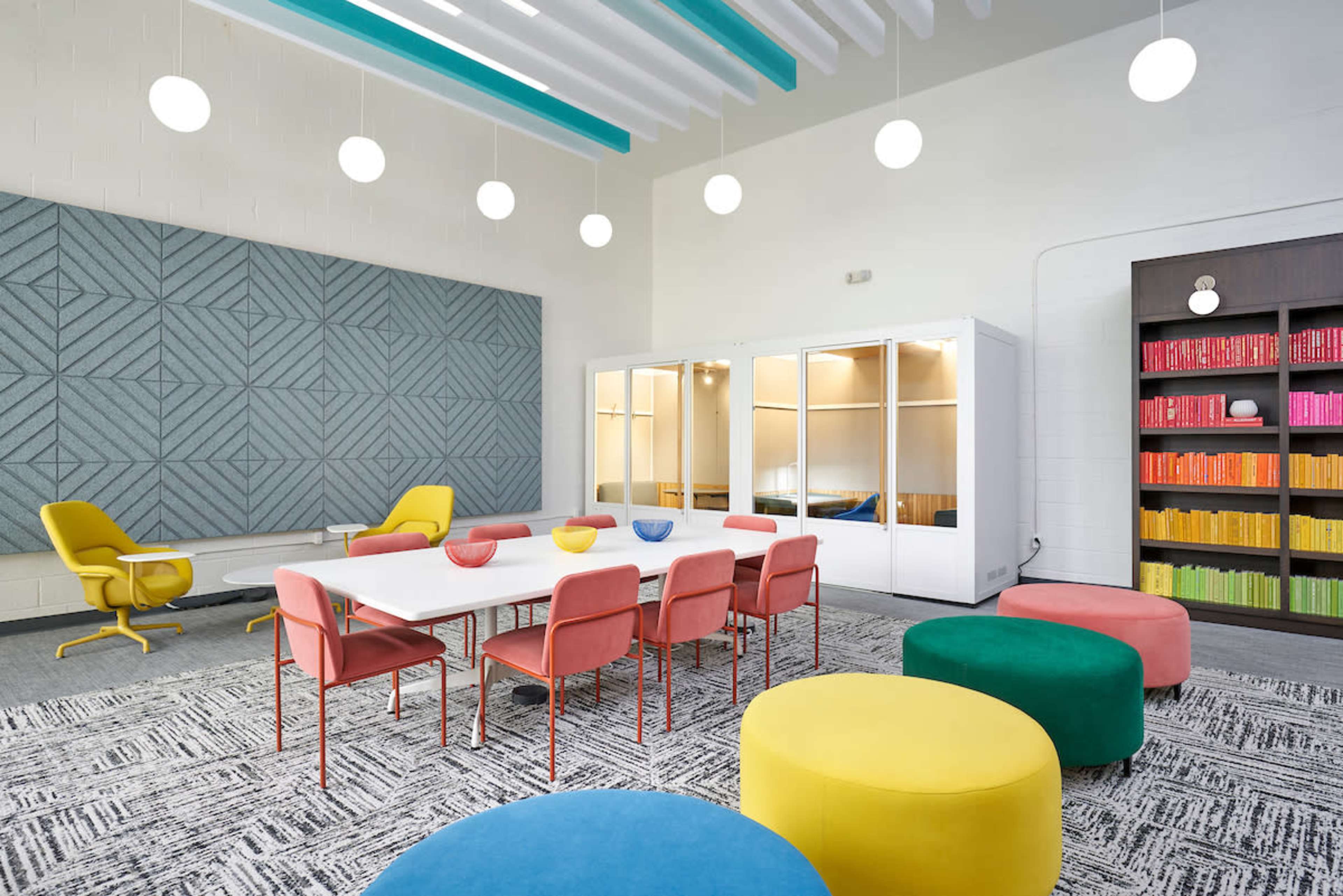 A bright meeting room features a white table surrounded by colorful chairs and stools, with a wall of books and a textured gray panel.