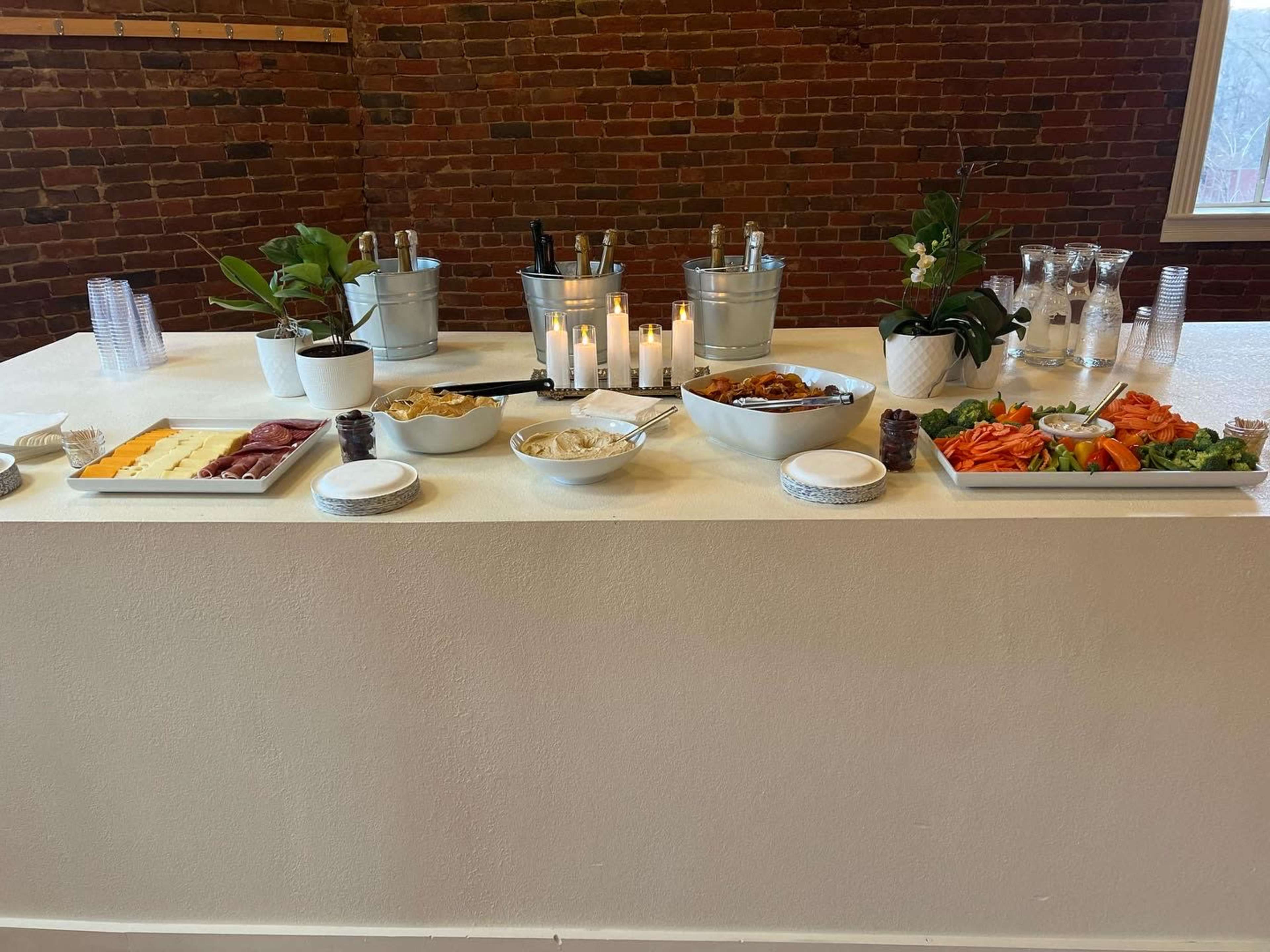 A food spread is arranged on a white table with platters of cheese, charcuterie, vegetables, and chips, alongside drinks in ice buckets and candles.