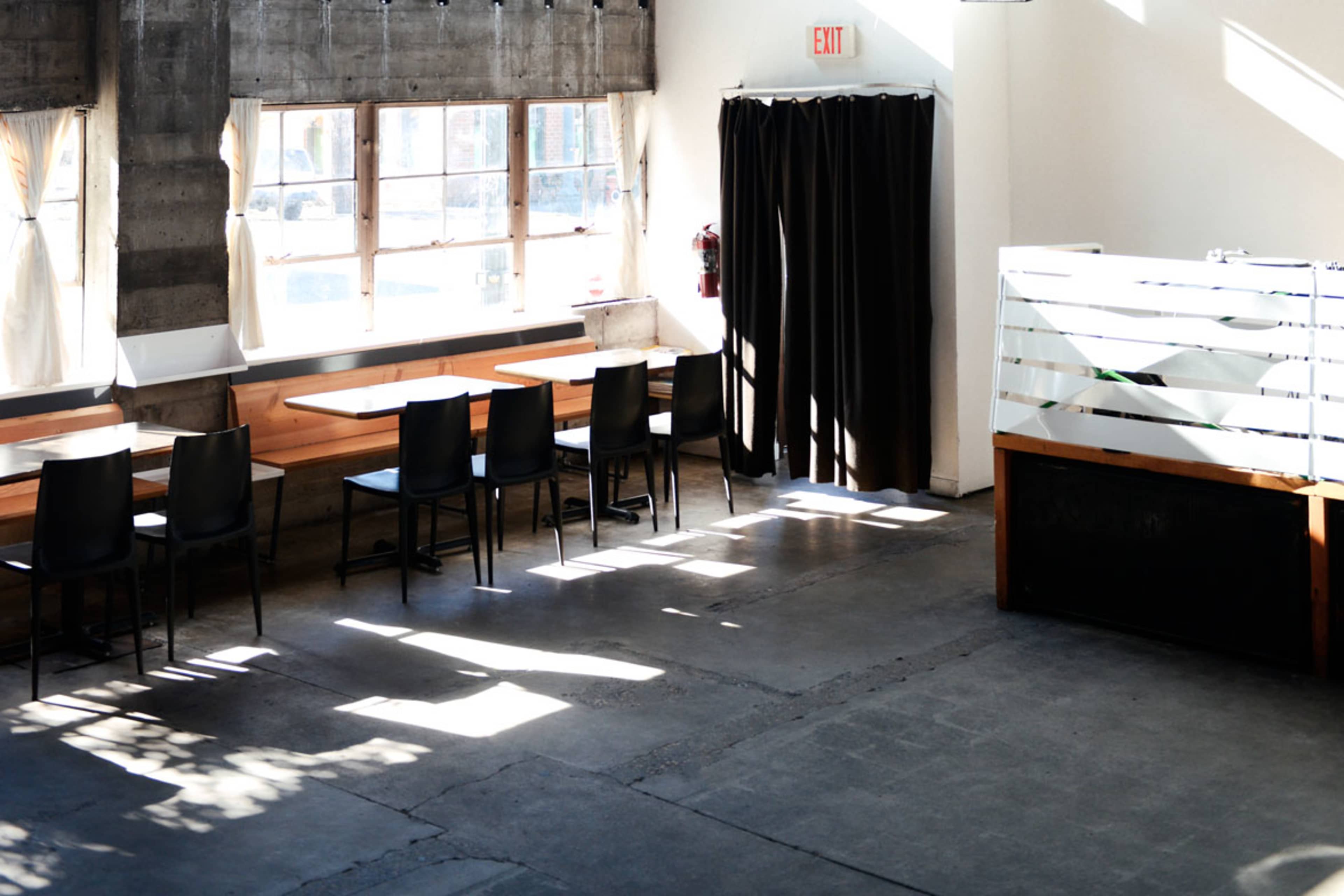 A minimalistic interior of a cafe with several empty black chairs and tables arranged near large windows, and a partition dividing the space.