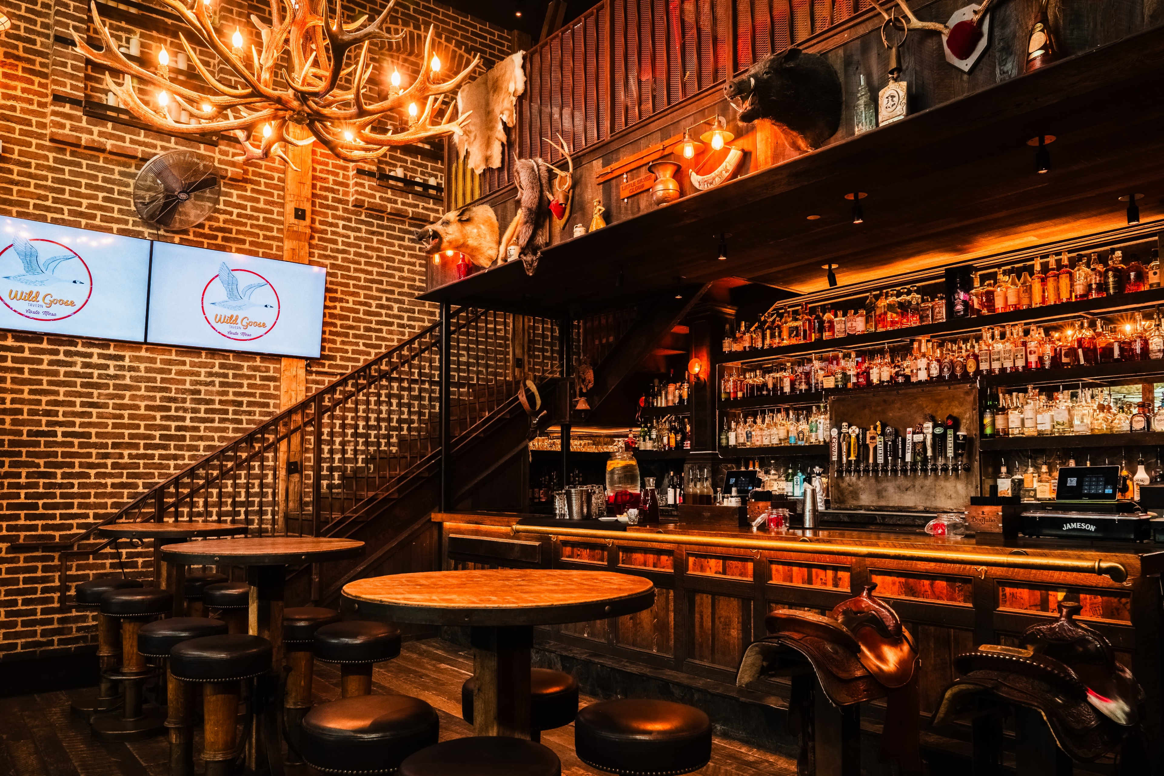 The image shows a rustic bar with a wooden counter, several round tables, and a staircase, surrounded by brick walls and adorned with antler decor and a large chandelier.