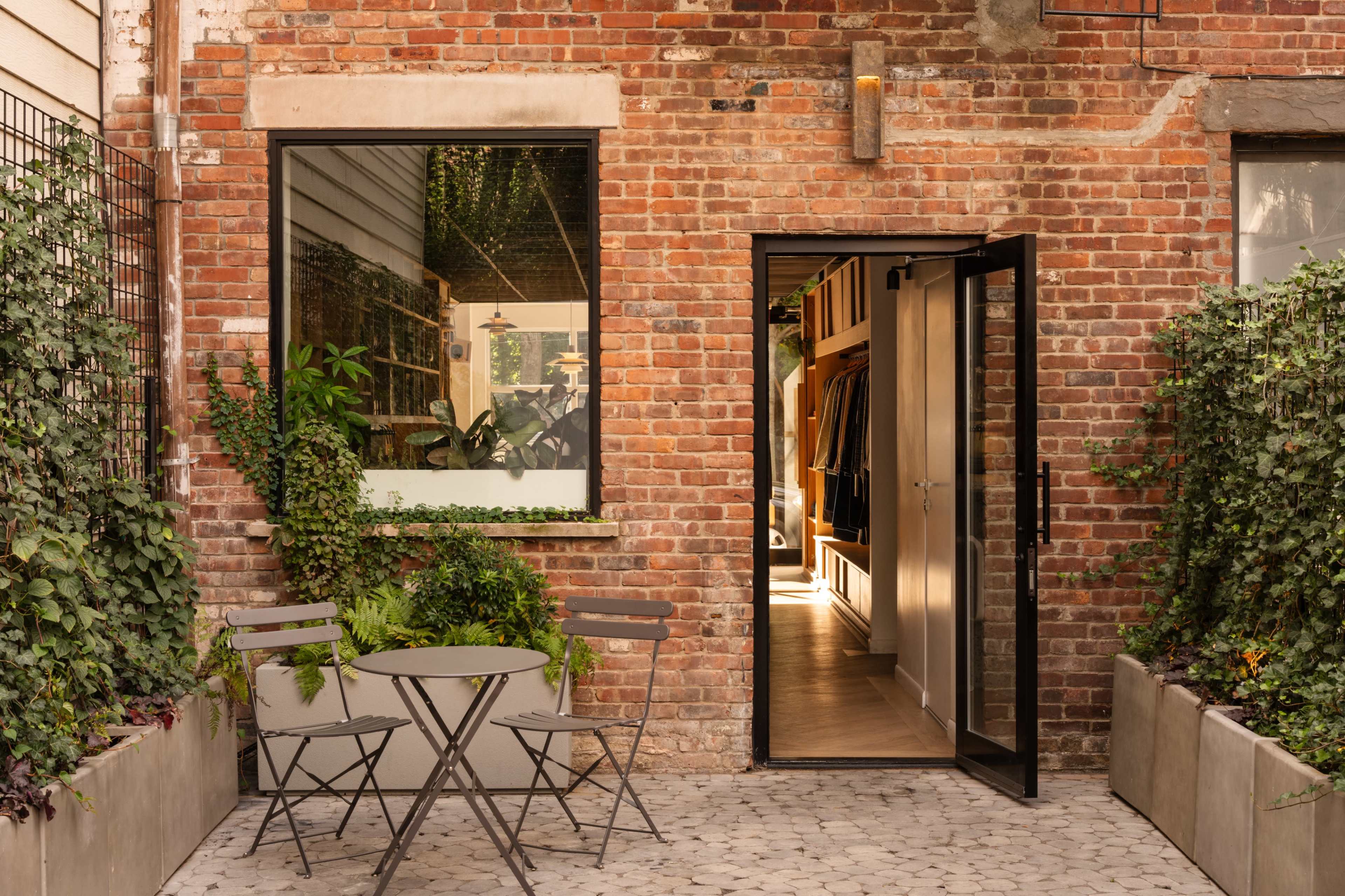 A brick wall surrounds an open doorway leading into a boutique, with a small table and chairs set up outside on a patterned stone surface.