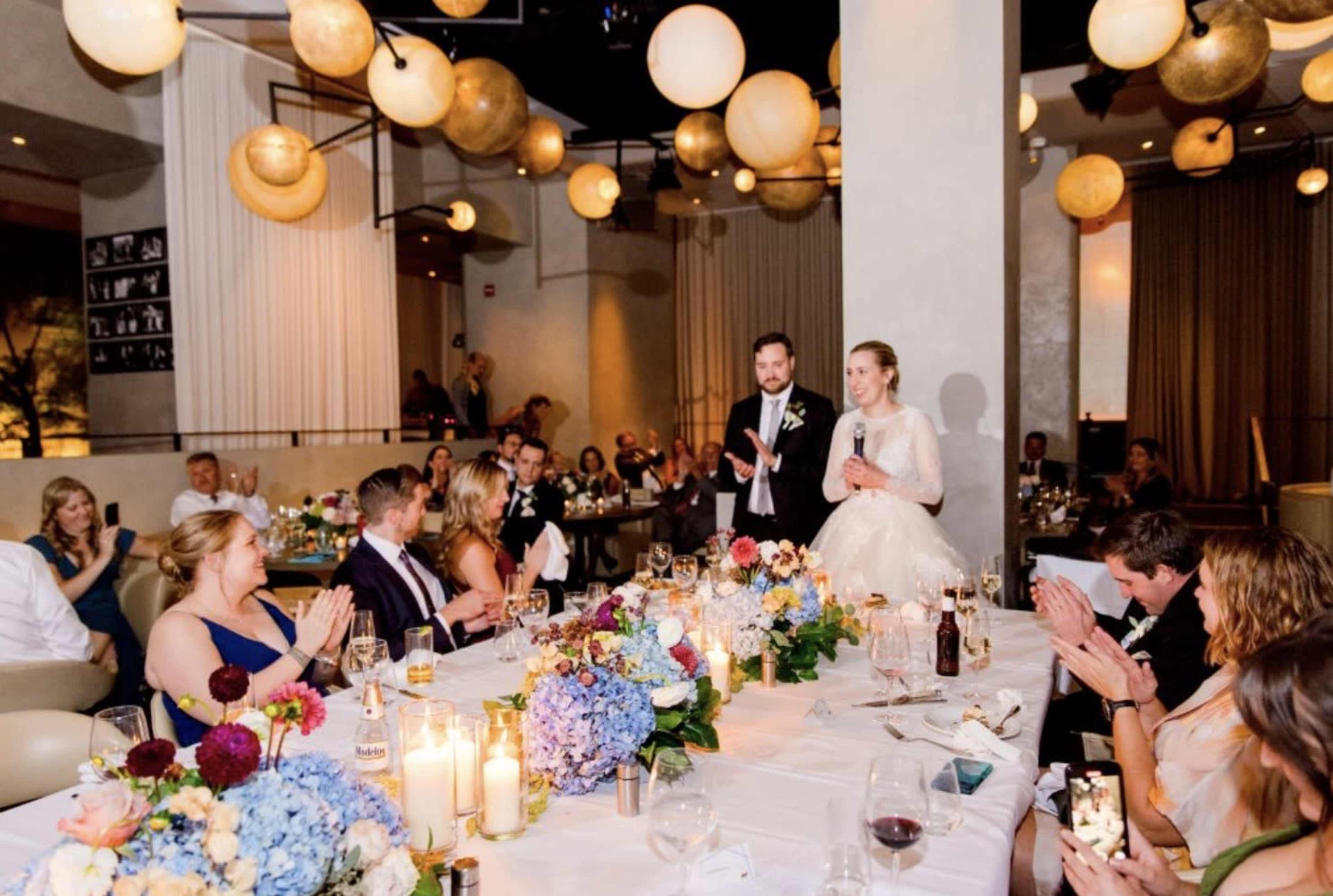 A bride and groom give a speech at a wedding reception surrounded by guests sitting at elegantly decorated tables.