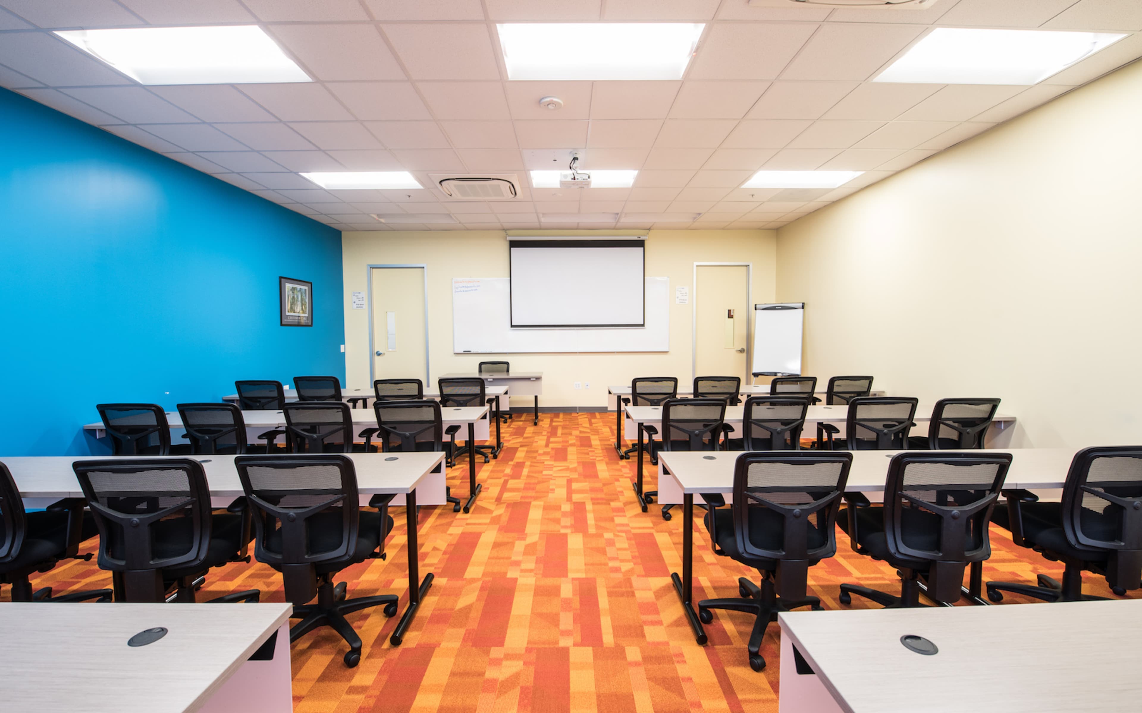 The image shows a classroom or meeting room arranged with rows of black office chairs facing a large projection screen and whiteboard in a brightly colored environment.
