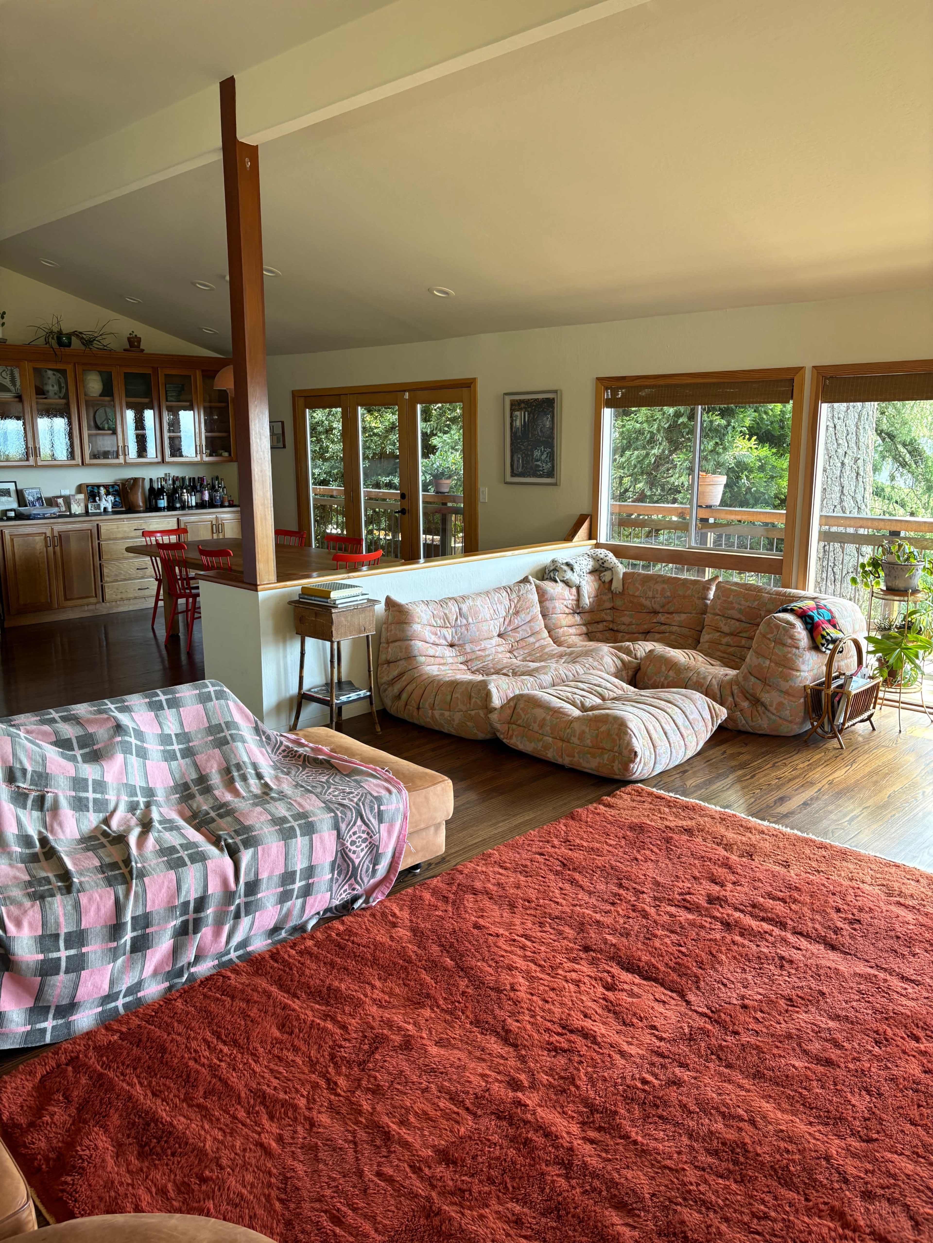 The image shows a spacious living area with a large, informal seating arrangement, a wooden dining area, and large windows overlooking greenery.