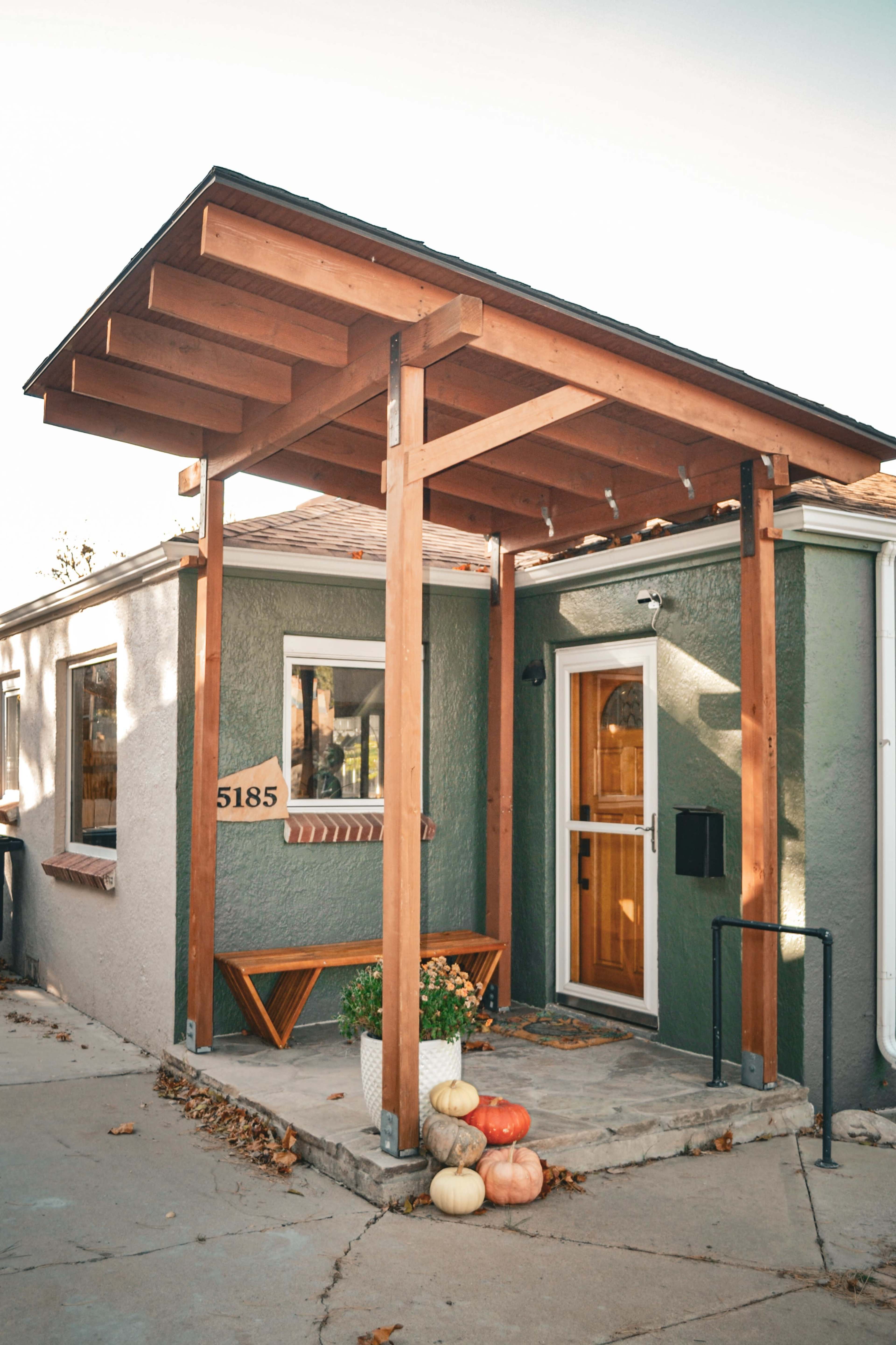 The image shows a small house entrance with a wooden overhang, a door, and several pumpkins on the porch.