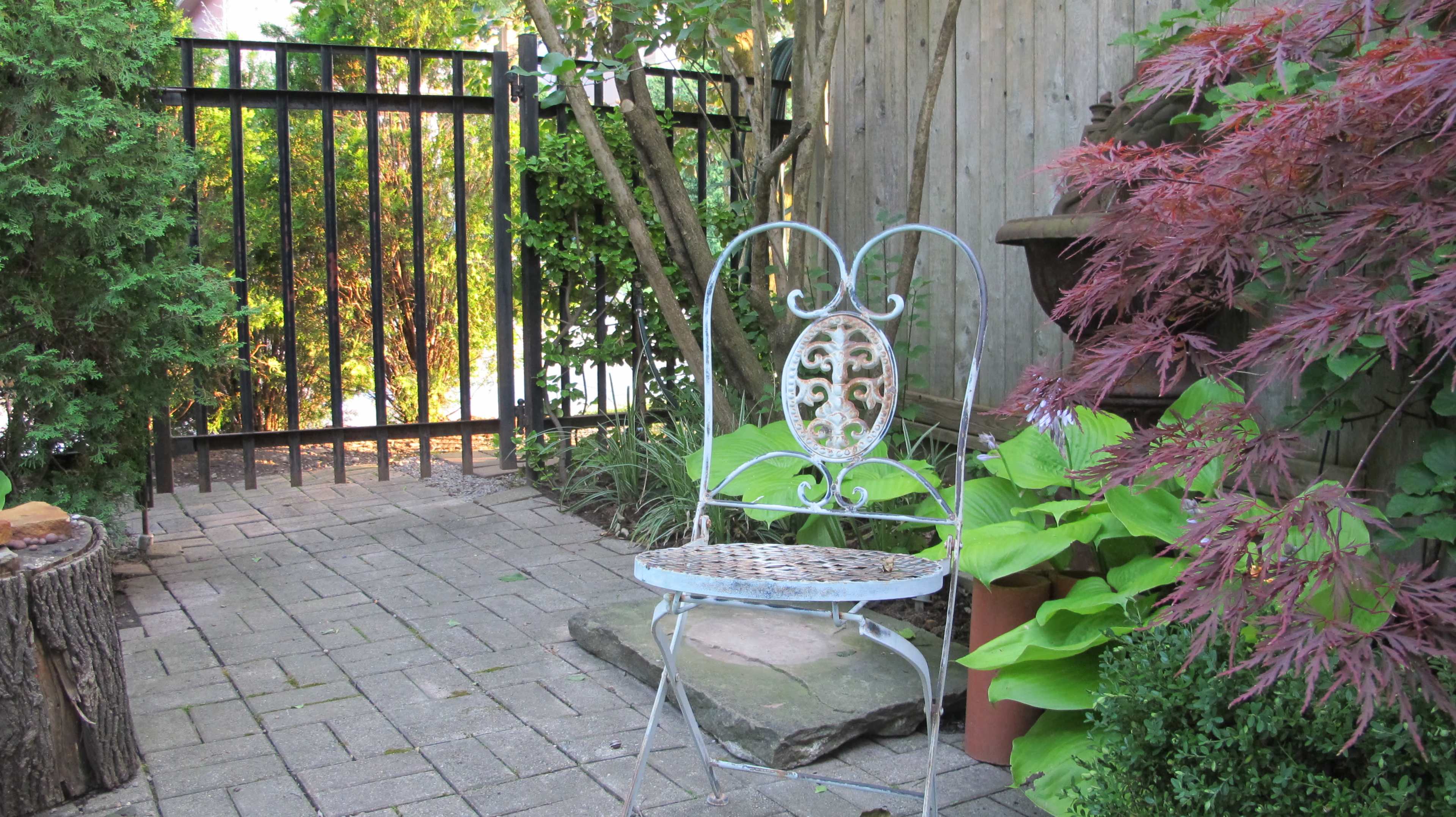 A wrought iron chair sits on a stone path beside lush green plants and a wooden fence gate.