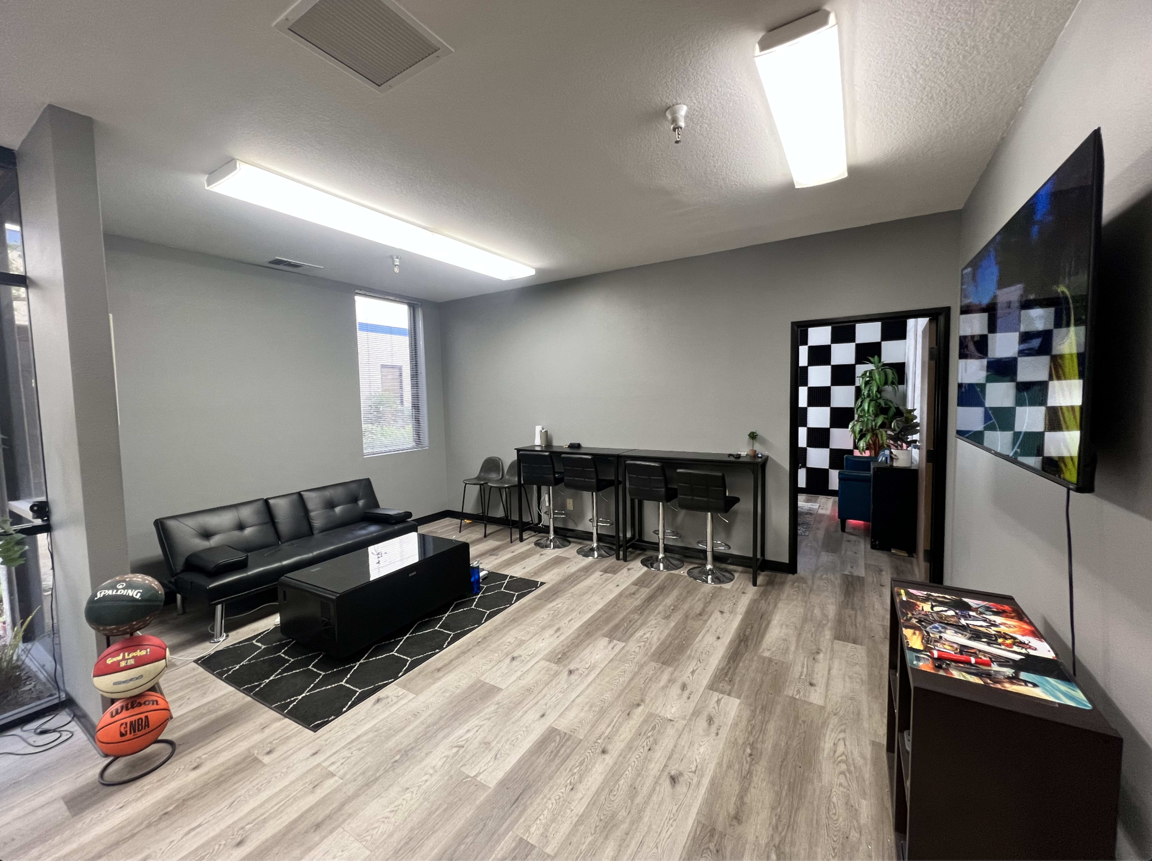 A modern lounge area with a black sofa, barstools along a counter, a large TV on the wall, and a patterned floor.