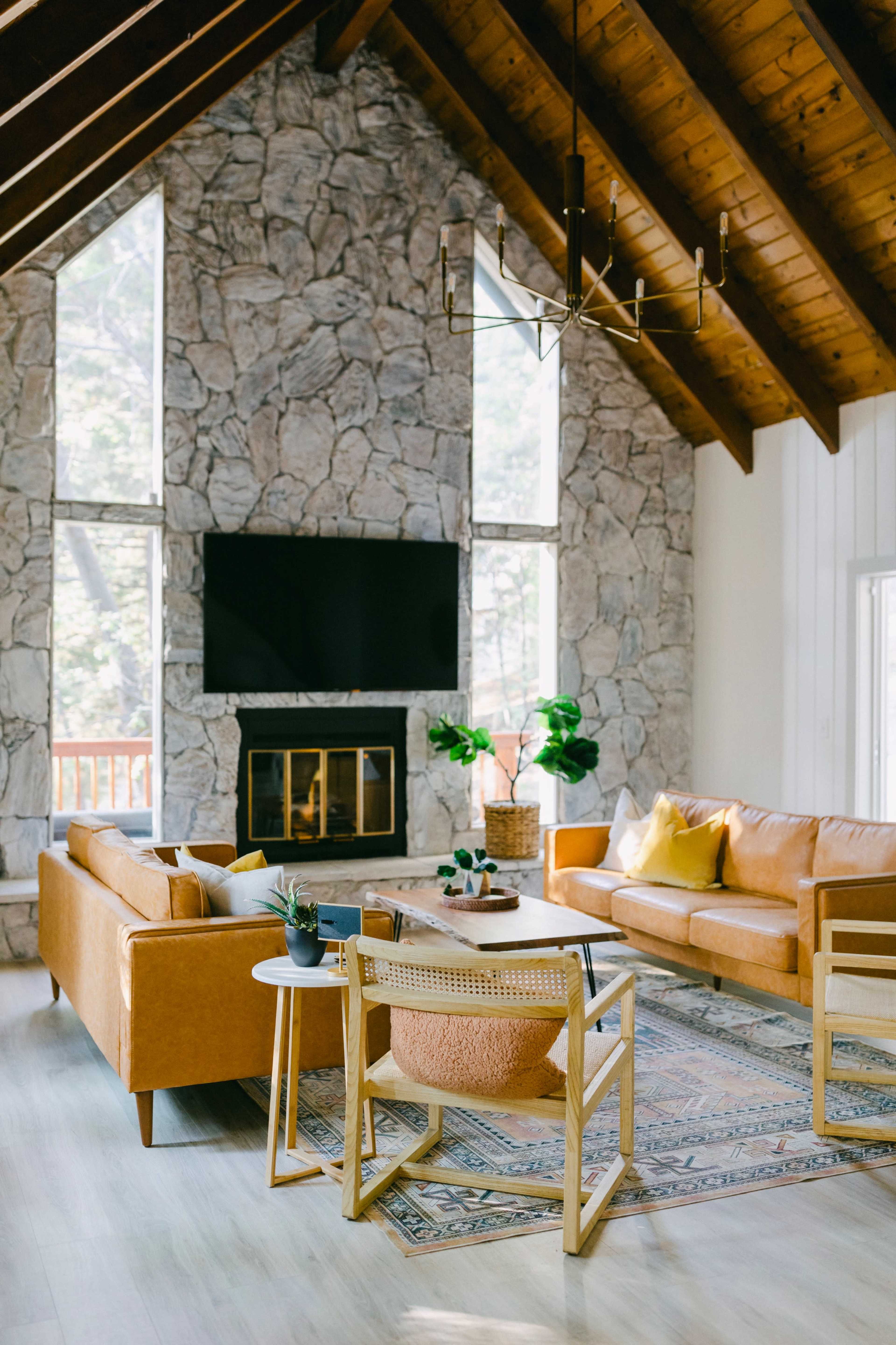 The living room features tall stone walls, a large window, a flat-screen TV above a fireplace, and modern furniture arranged around a patterned rug.