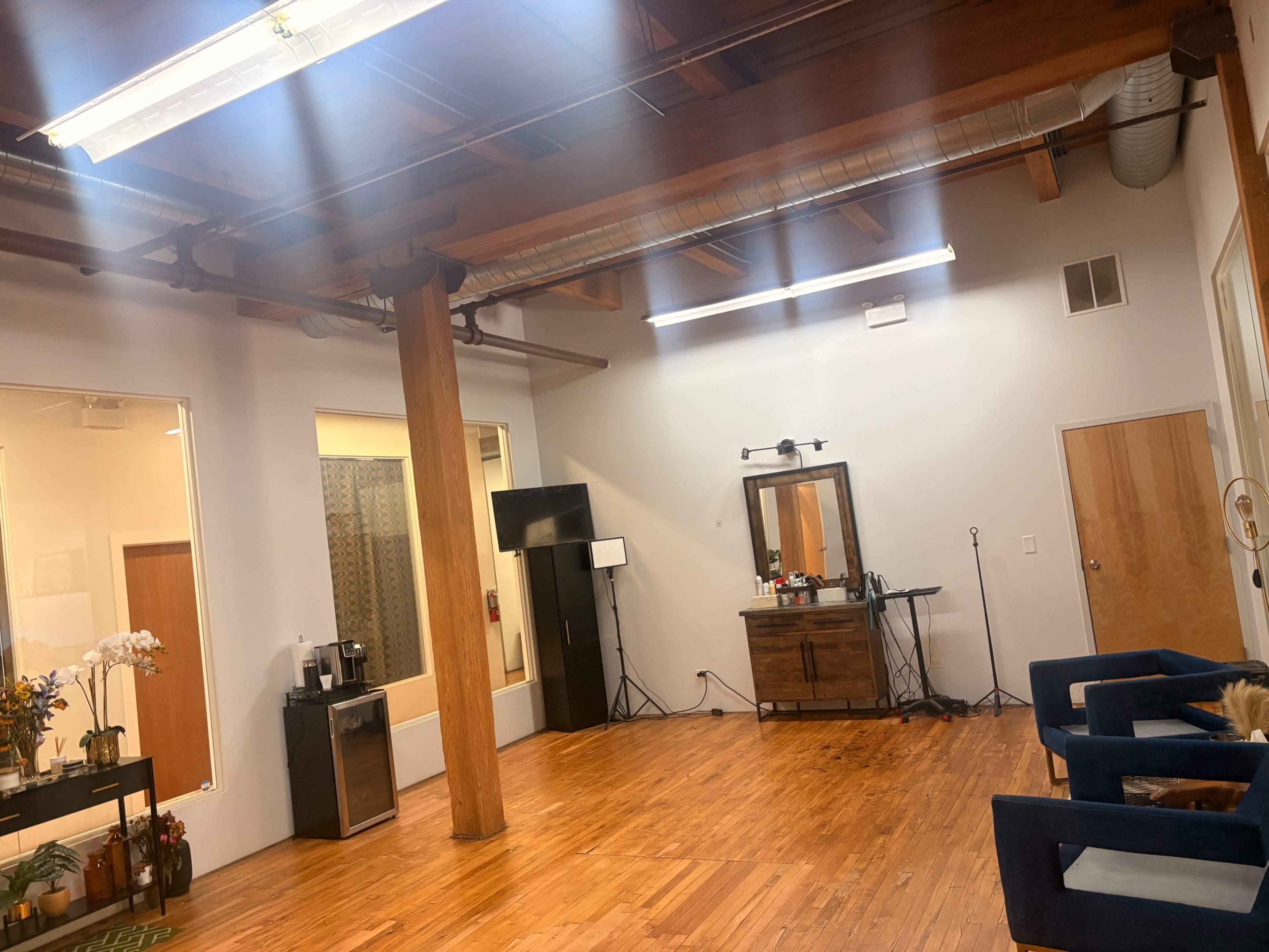 The image shows a spacious and well-lit room with wooden floors, exposed beams, a mirrored vanity area, and a few pieces of furniture, including a cabinet and blue chairs.