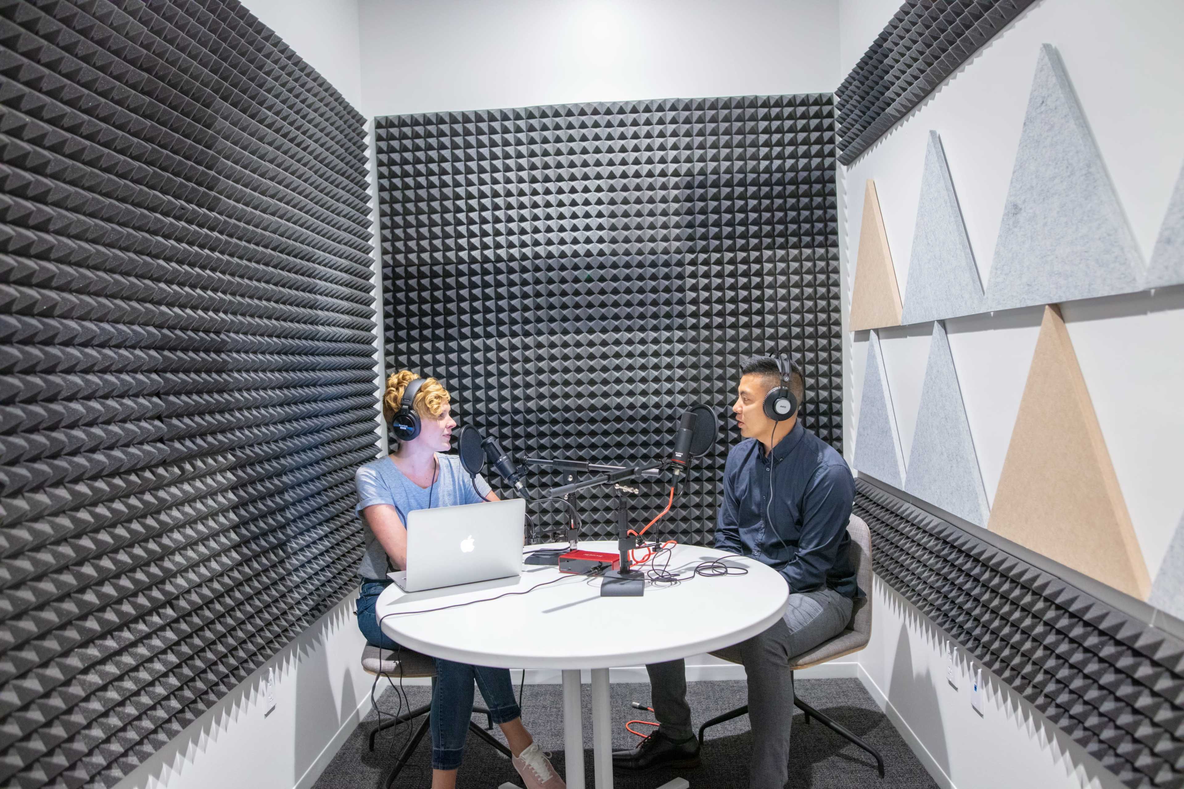 A woman and a man sit at a round table in a soundproof podcast studio, each wearing headphones and speaking into microphones.