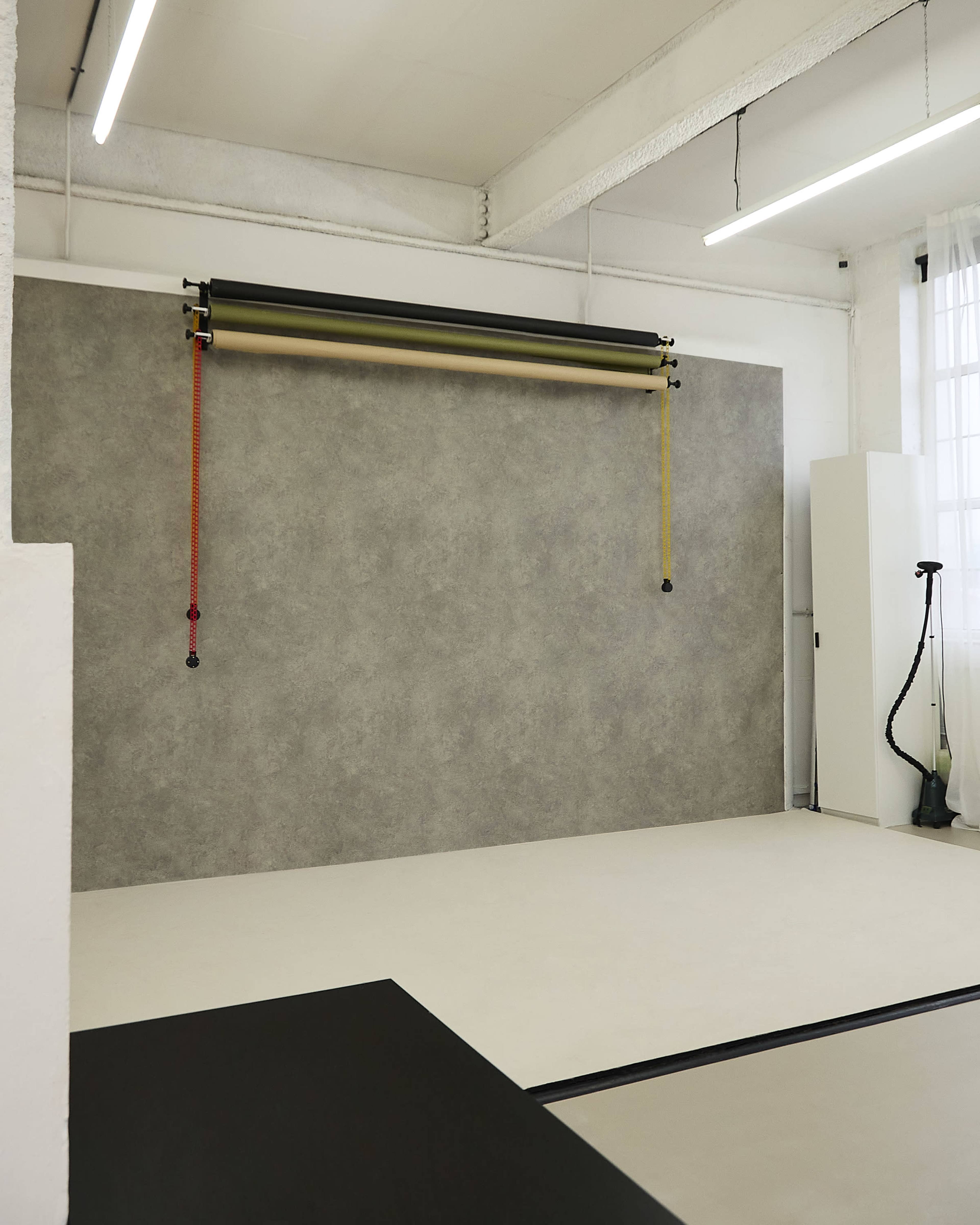 The image shows a minimalist photography studio with a textured backdrop on the wall, a plain floor, and a green garment steamer in the corner.