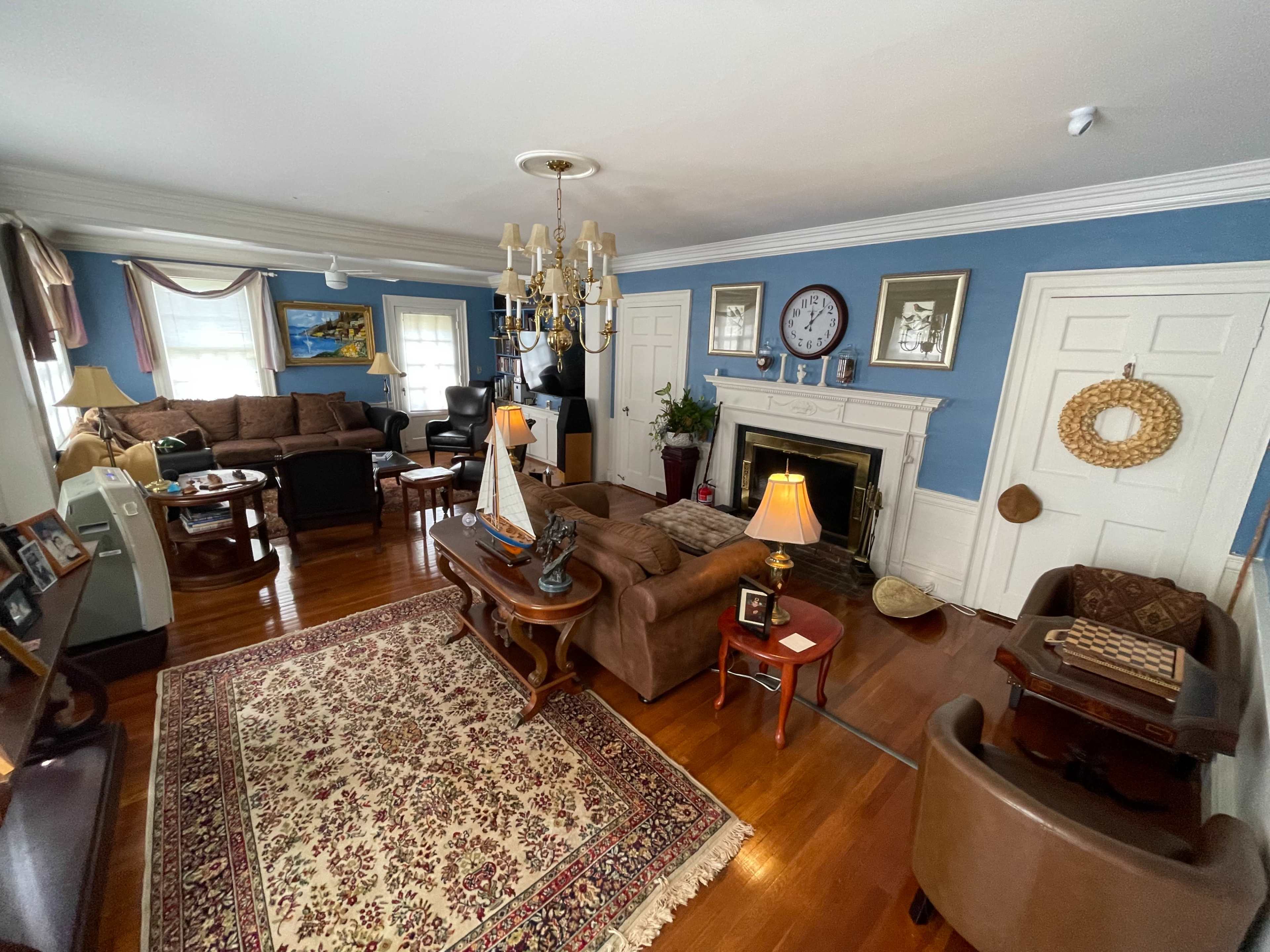 The image shows a cozy living room with a fireplace, a chandelier, and various seating areas arranged around a patterned rug.
