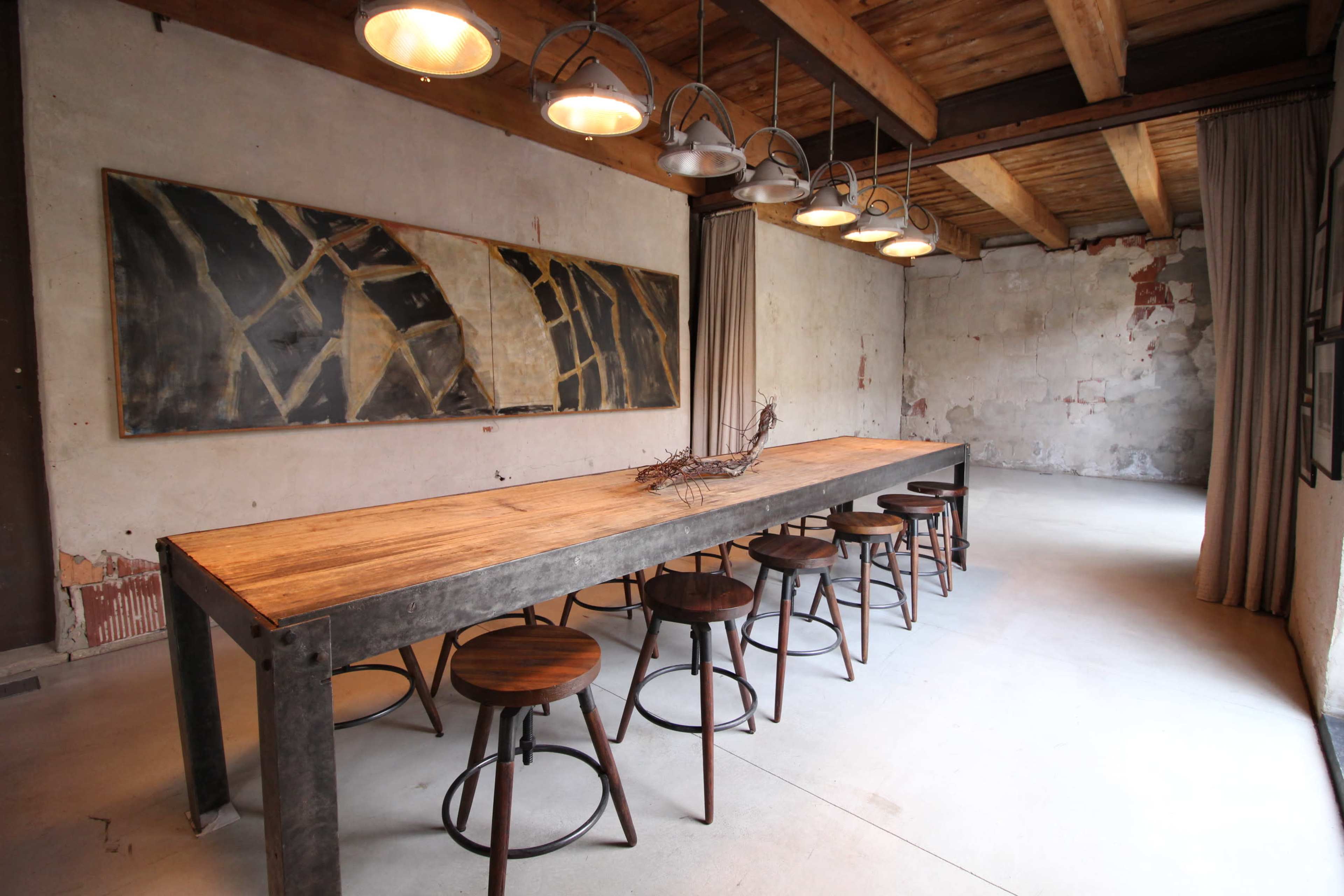 A long wooden table with metal legs is surrounded by several stools in a modern, industrial-style dining room with exposed brick walls and overhead lighting.
