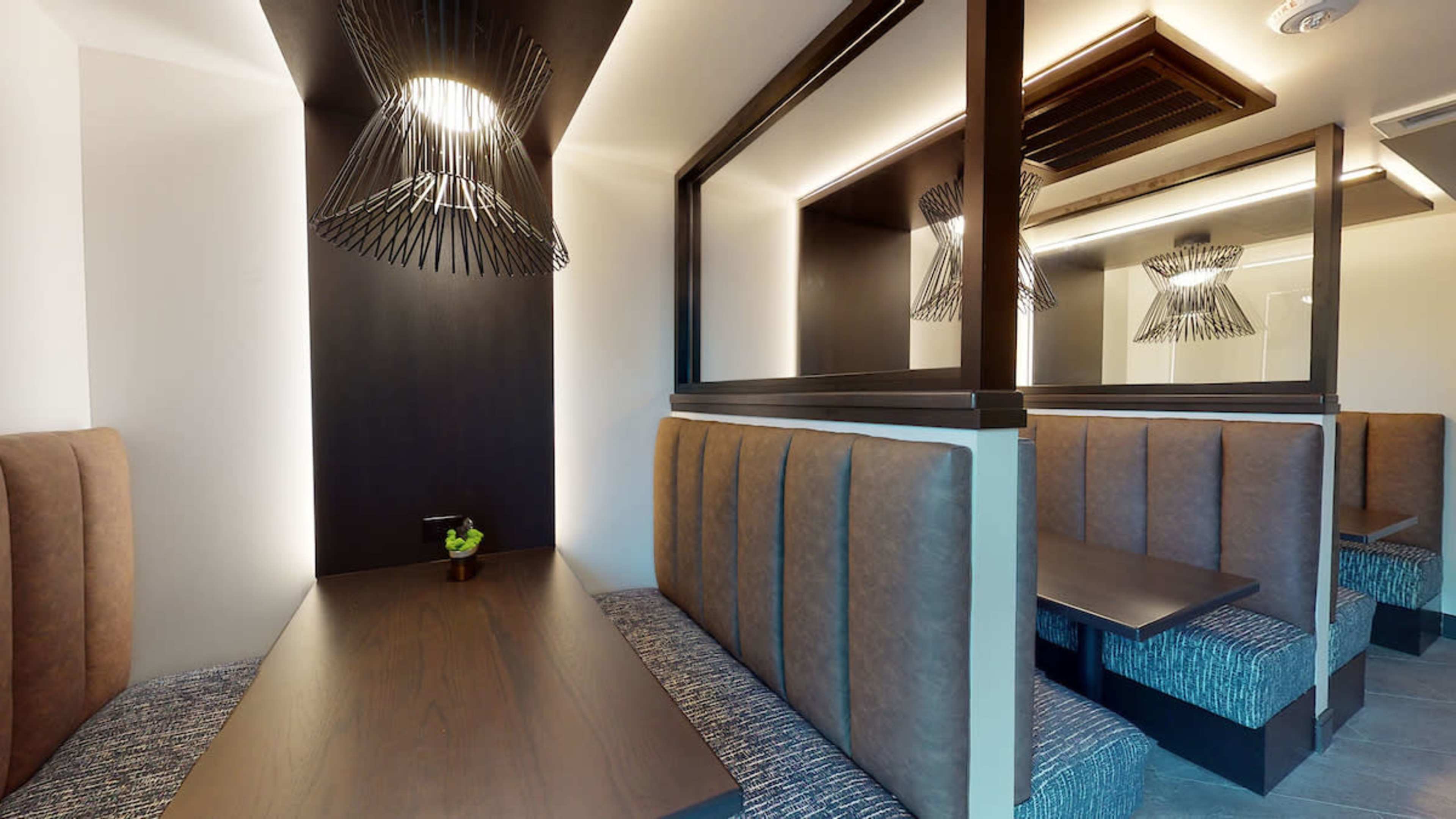 The image shows a modern dining space with cushioned benches and wooden tables, featuring pendant lights and mirrored walls.