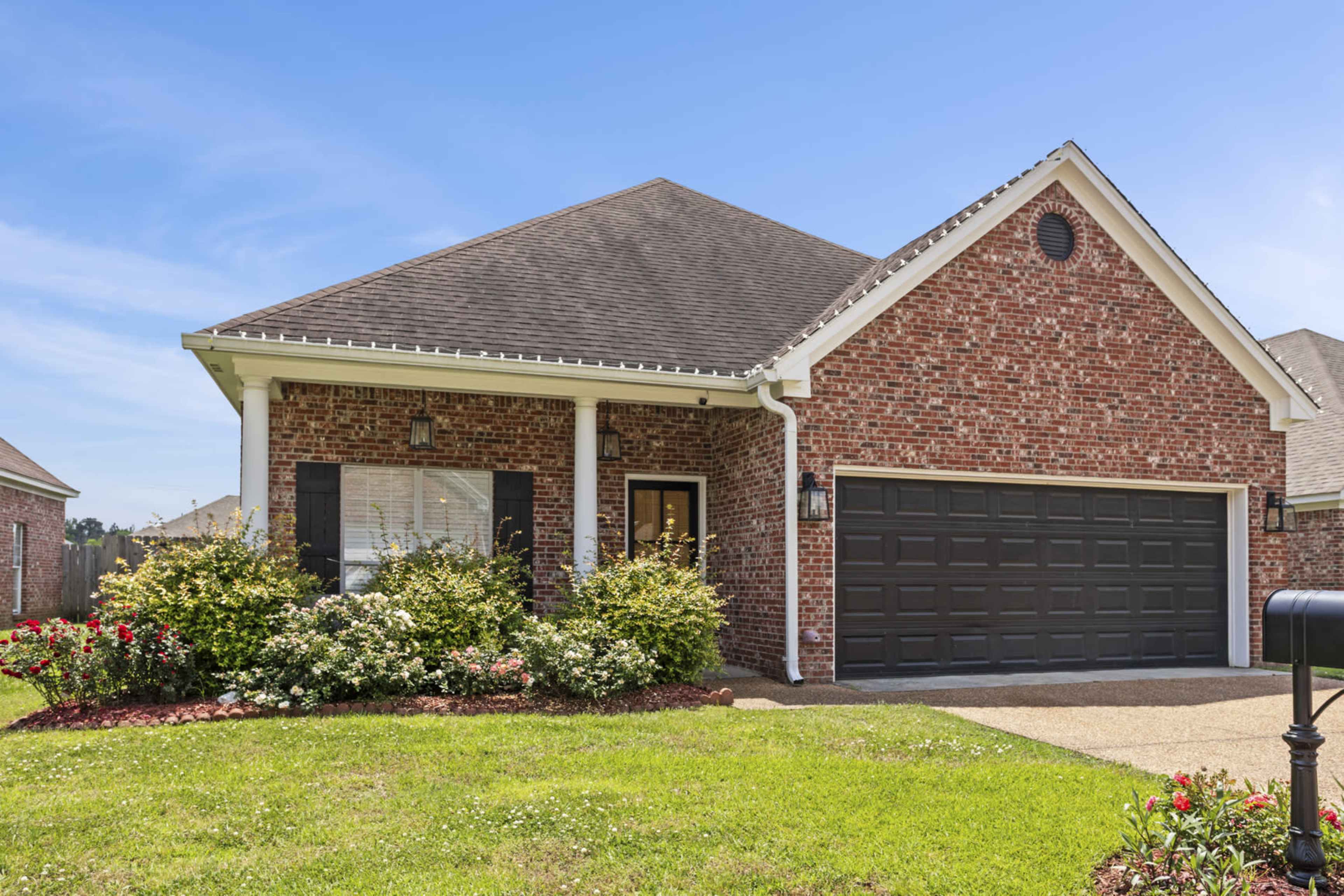 A single-story brick house with a gabled roof and a dark garage door is surrounded by green grass and flower bushes.