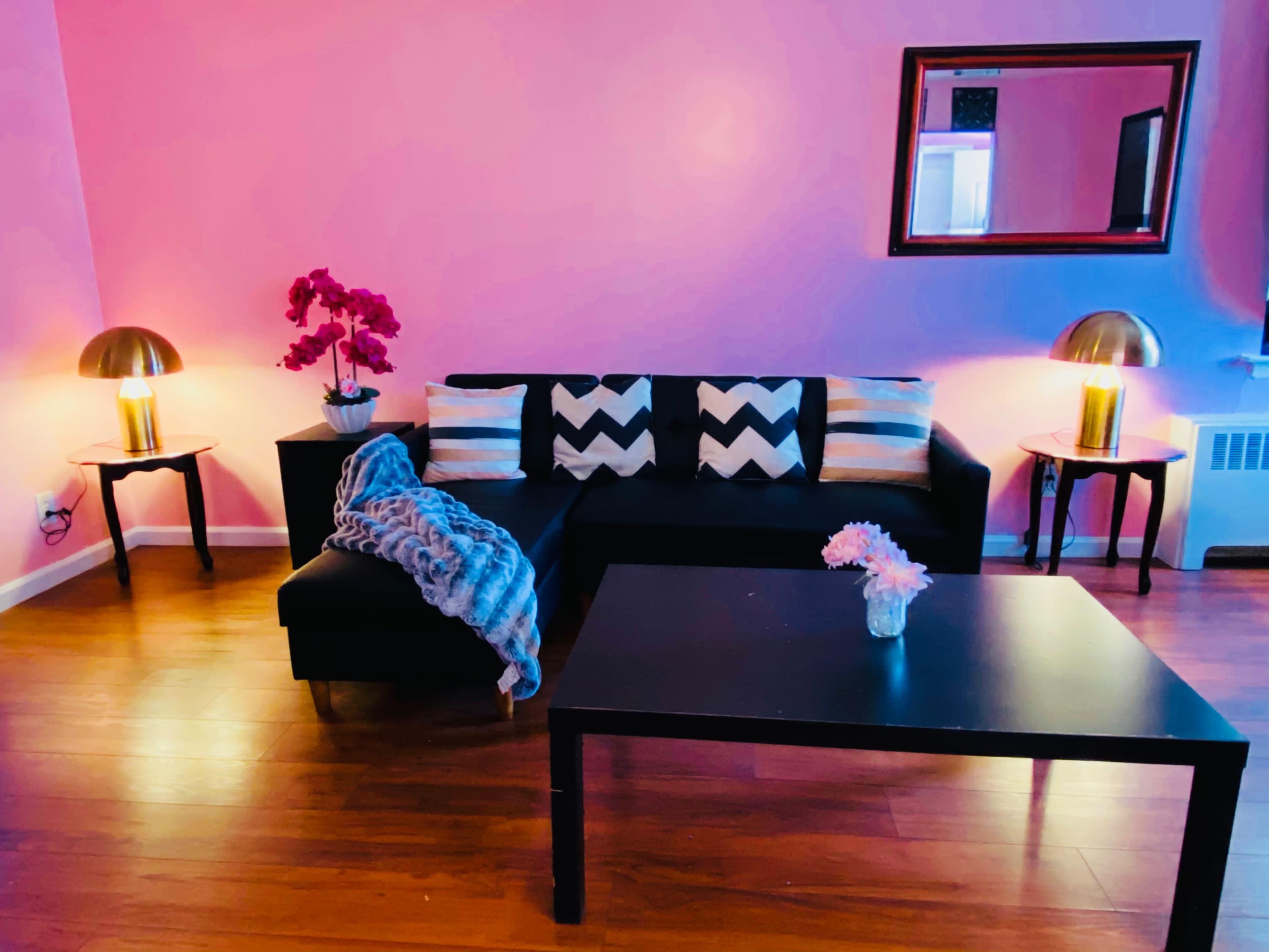 A modern living room with a black sectional sofa, decorative pillows, a coffee table, and two lamps, all set against pink walls.