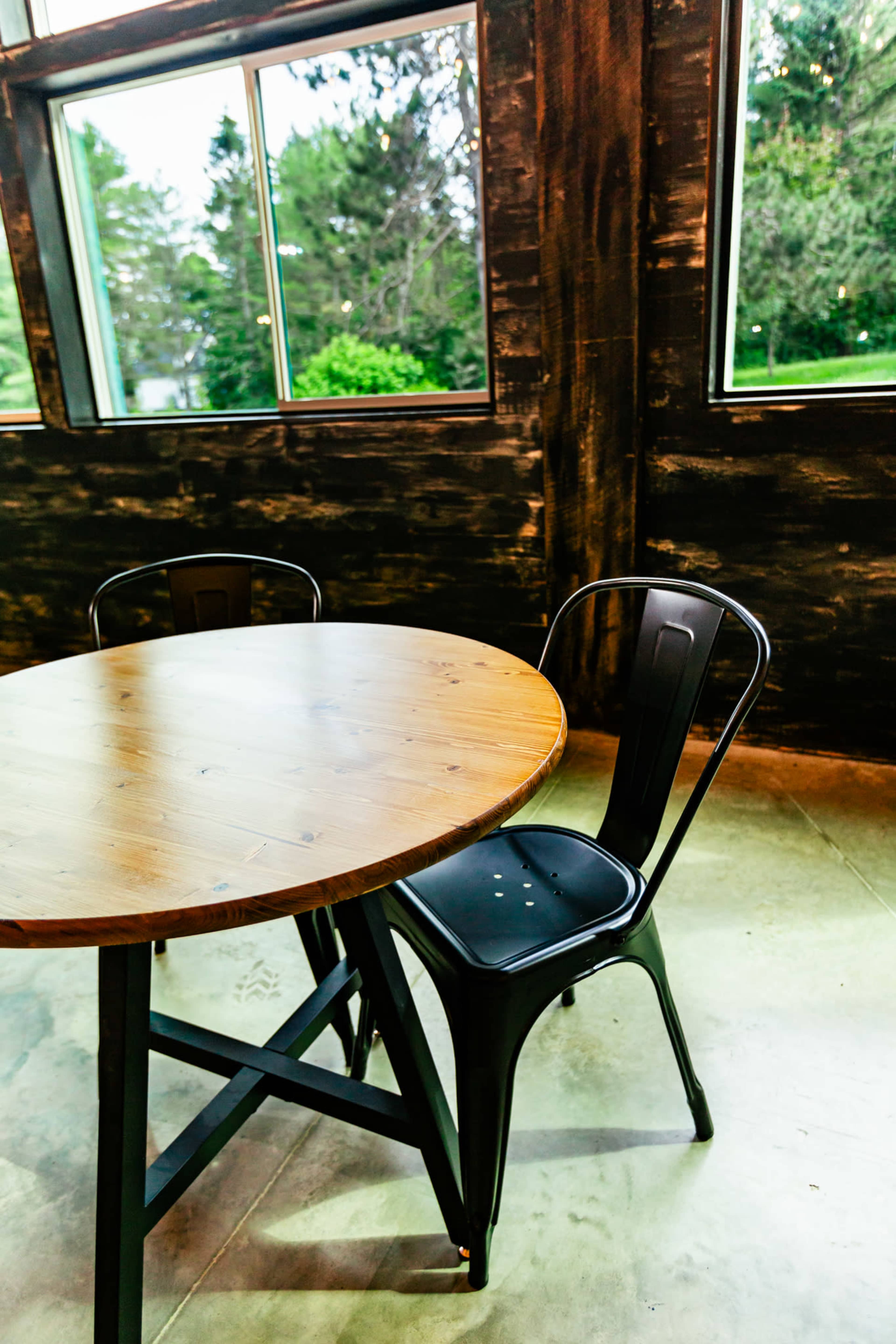 A round wooden table with a metal base stands beside a black metal chair in a room with large windows overlooking greenery.