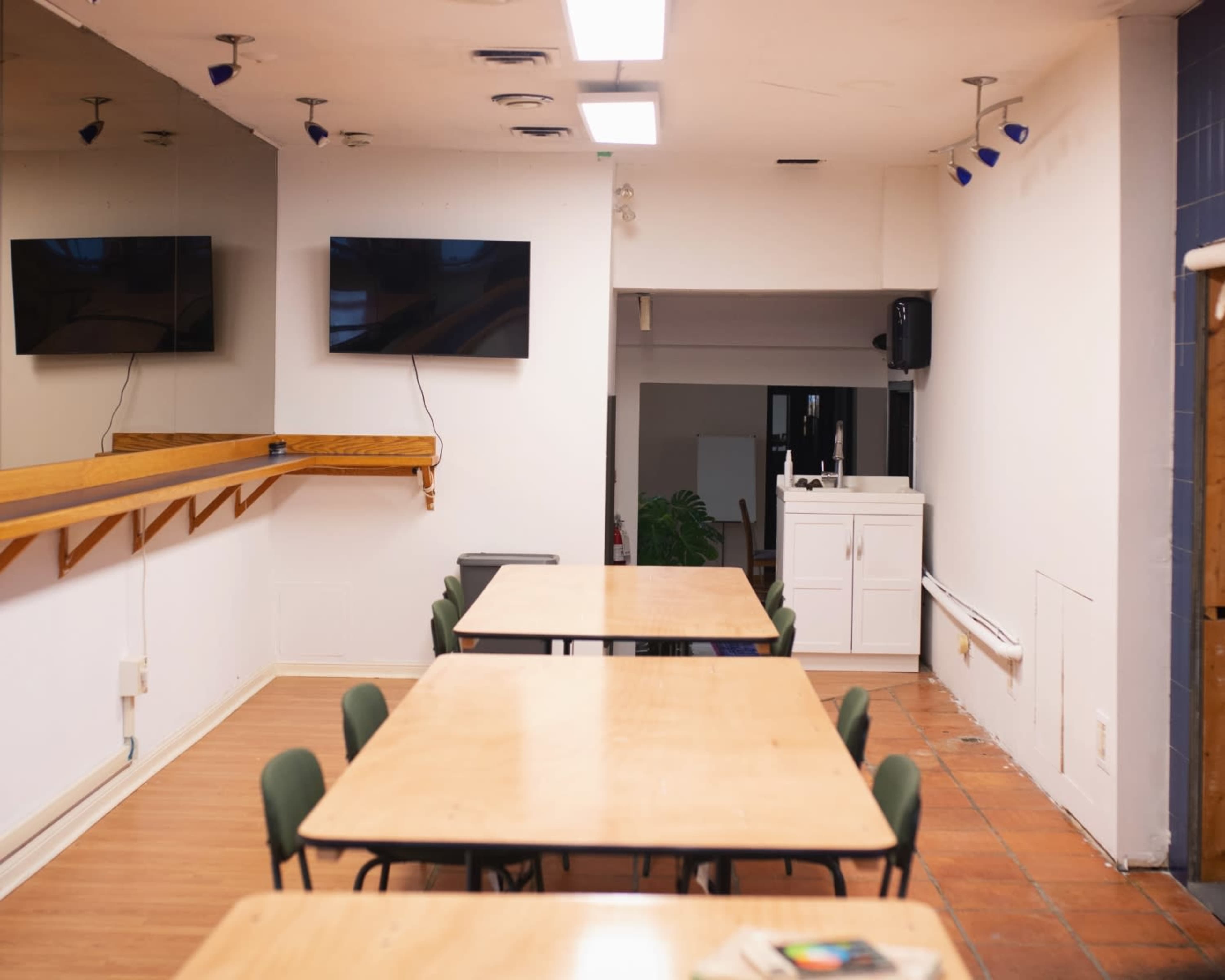 The image shows a large, empty room with several wooden tables arranged in rows and two mounted televisions on the wall.