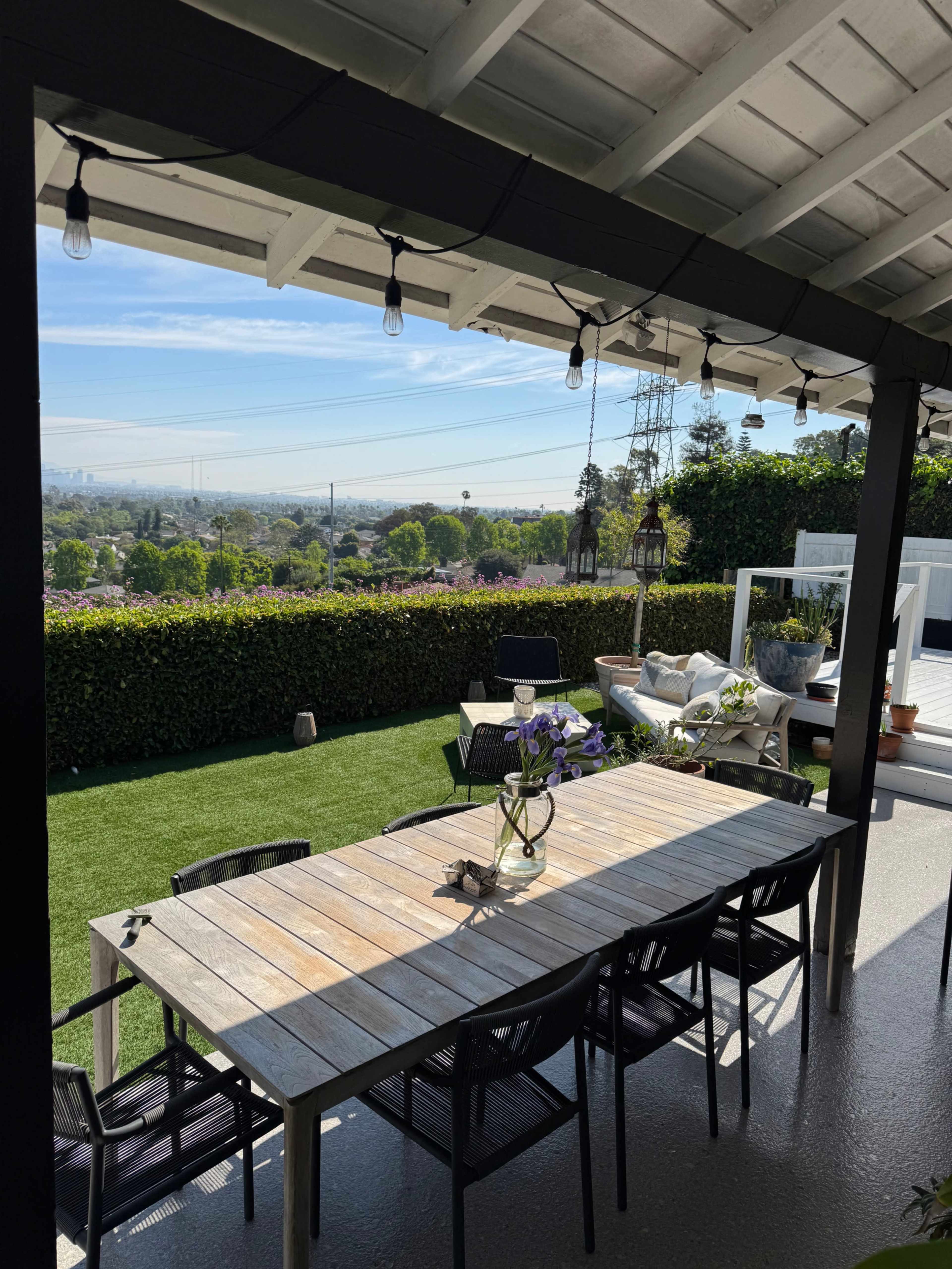A wooden dining table with black chairs is set on a patio overlooking a landscaped garden and cityscape in the background.